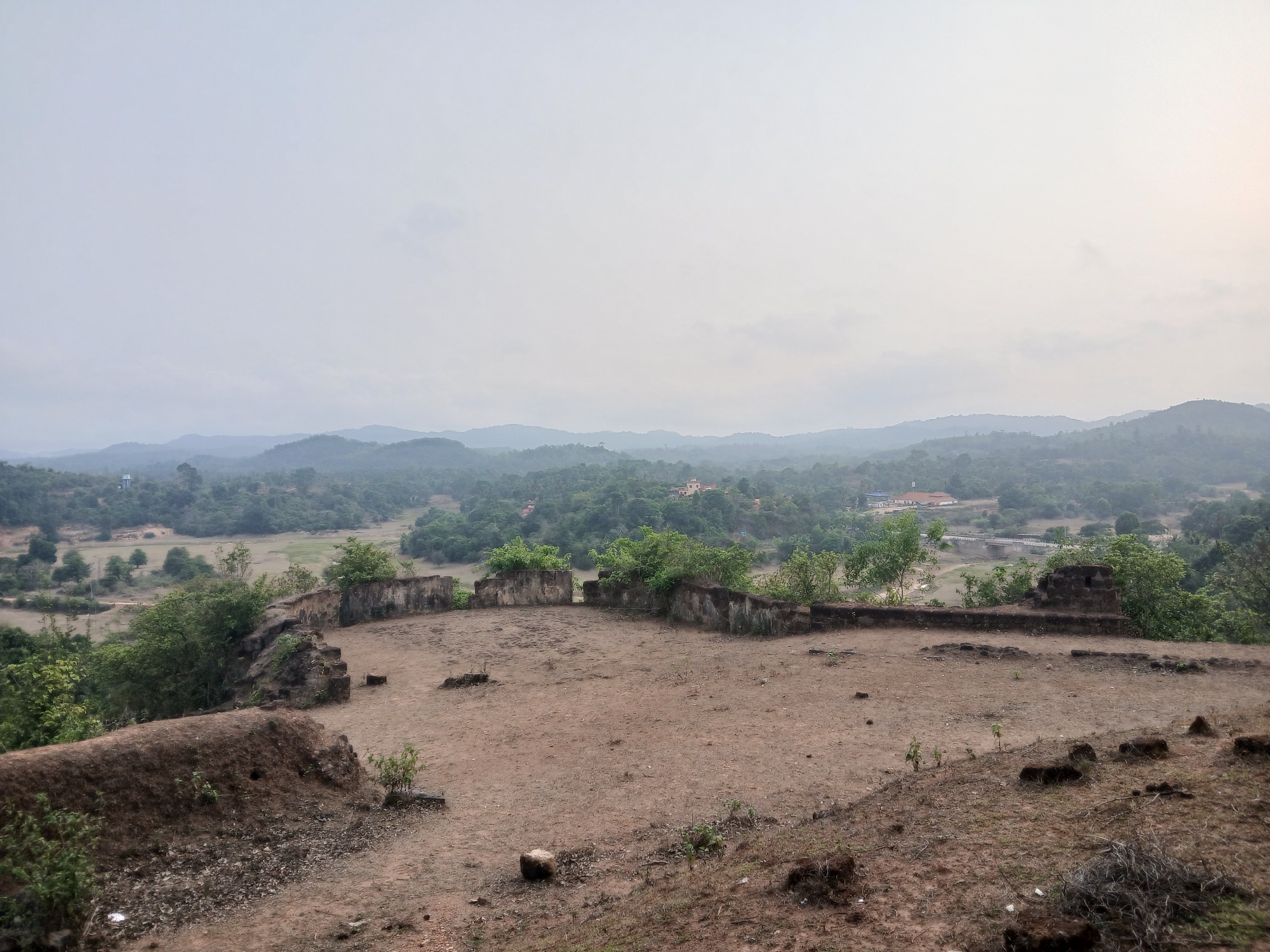 The image depicts the ruins of Nagara Fort, located in the village of Nagara, India. The foreground shows an earthen and stone structure, possibly a remnant of a wall or fortification. The ground is barren and appears dry. Behind this structure, there is a partially intact wall or enclosure, overgrown with vegetation. The background reveals a landscape of rolling hills and dense green forests, shrouded in a hazy atmosphere. A few buildings are visible in the distance, partially obscured by the vegetation and mist. The sky is overcast, contributing to the overall dull and muted color palette of the scene.