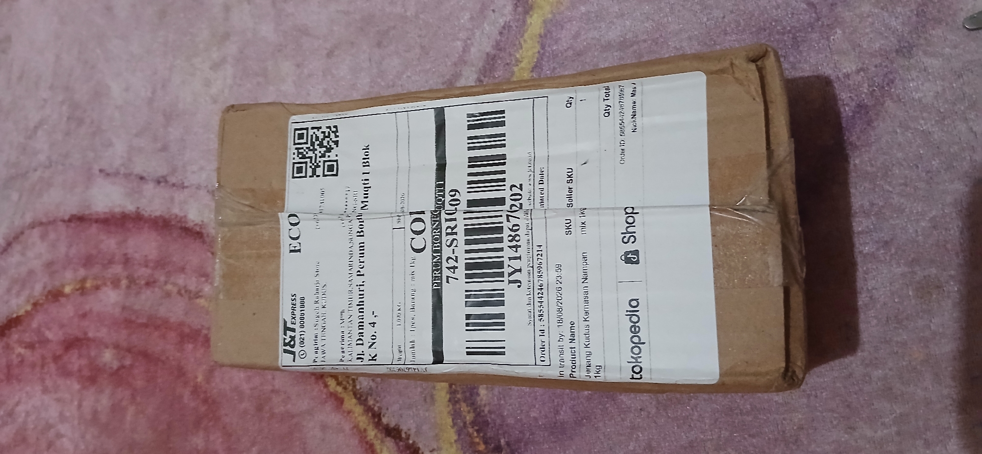 The image shows a brown cardboard box with shipping labels. The labels indicate that the package is being sent via J&T Express, a courier service. One label has the text "ECO" and a QR code. Another label contains shipping information, including sender and receiver details. The receiver's address is listed as "Jl. Damanhuri, Perum Bornet Muqti 1 Blok K No. 4,-". This indicates the package is destined for Samarinda, Indonesia, as Jl. Damanhuri is a well-known street in the city. The box is placed on a textured surface that appears to be a blanket or rug, predominantly in shades of pink and red. The lighting suggests the image was taken indoors, possibly at night.