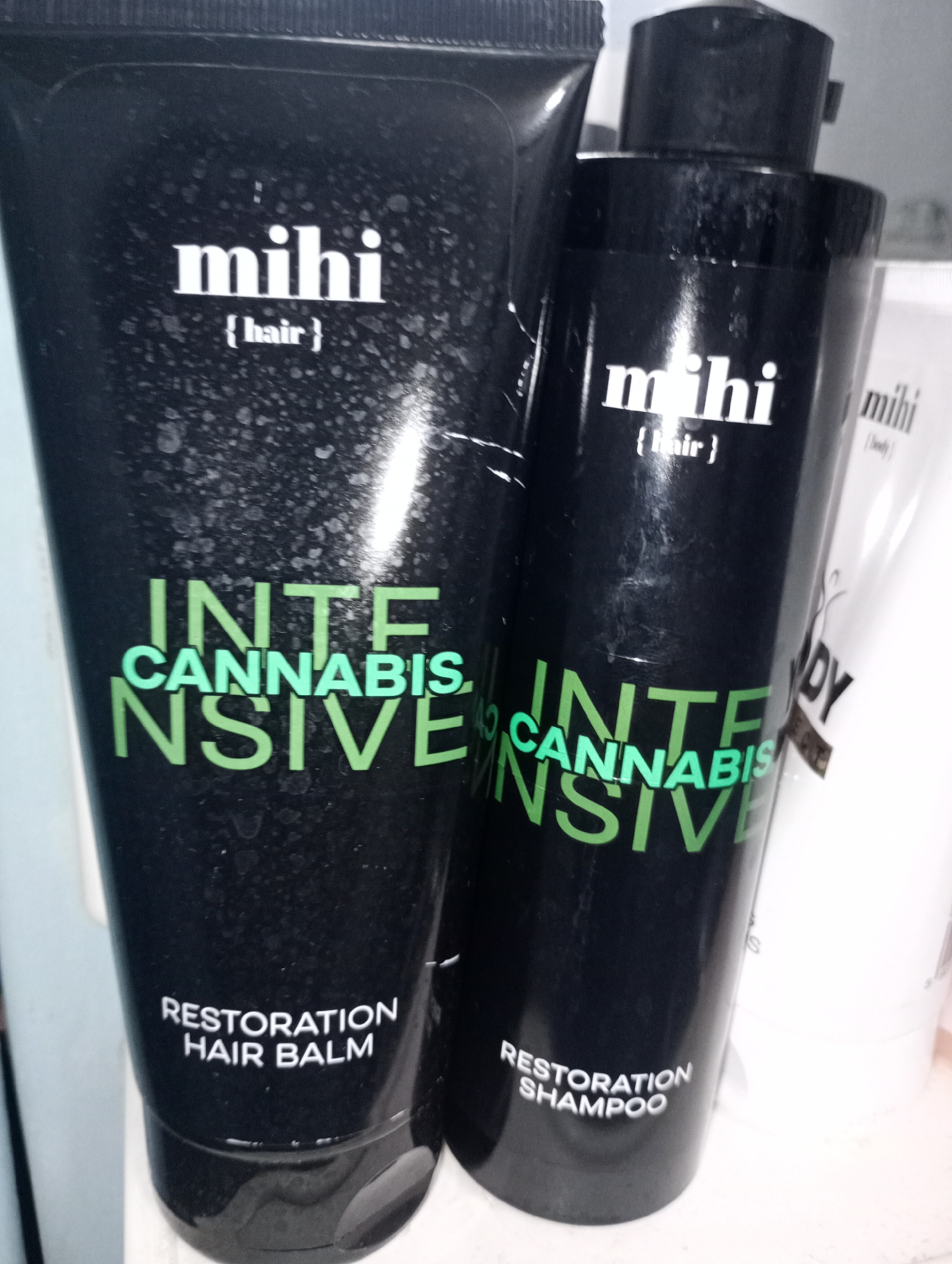 The image shows two black bottles of hair products. The bottle on the left is a tube labeled "mihi {hair} INTENSIVE CANNABIS RESTORATION HAIR BALM". The bottle on the right is a pump bottle labeled "mihi {hair} INTENSIVE CANNABIS RESTORATION SHAMPOO". There is a white bottle partially visible behind them, also with "mihi" branding. The bottles are set against a light-colored background, possibly a shelf or counter. There are no people visible in the image. The lighting is somewhat dim, with some glare on the bottles. The location of Harmanli, Bulgaria is not depicted or suggested in this image.