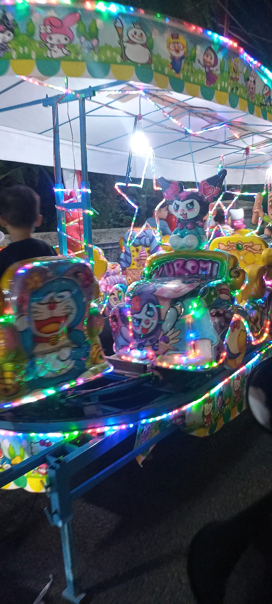 An amusement ride at night features bright, colorful lights and cartoon characters. The ride has a canopy decorated with images of characters such as My Melody, a snowman, and Doraemon. The seats on the ride are also decorated with characters, including Doraemon and My Melody's character Kuromi. A young child is seated in the ride, viewed from behind. The ride is positioned on a dark, outdoor surface, possibly asphalt. The scene is located in Kalimati, Indonesia.
