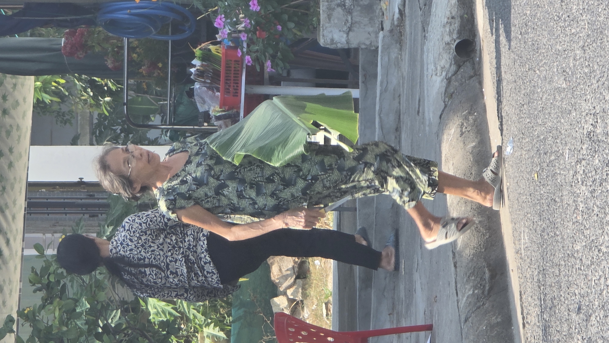 The image shows two women standing on a sidewalk near a road. The woman in the foreground, older with short gray hair and glasses, wears a green patterned top and capri pants, and sandals. She carries a large green leaf and a knife. The woman behind her wears a black and white patterned top and black pants, and sandals. The background includes greenery, a red plastic crate, and a blue hose. The location is possibly in or near Bắc Nha Trang, Vietnam, considering the vegetation, architectural style, and clothing. The women are likely involved in a domestic activity, possibly preparing food or materials.