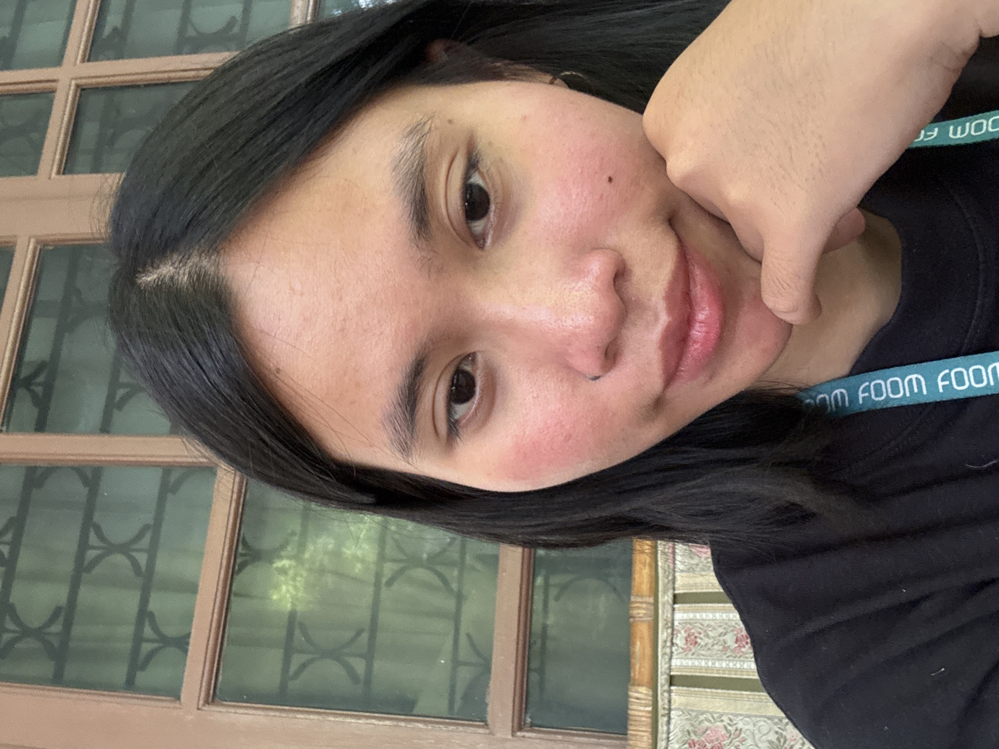 A young woman with long, dark hair and dark eyes is looking at the camera. She is resting her chin on her right hand. Her skin is light brown, and she has rosy cheeks. She is wearing a black t-shirt with a blue lanyard around her neck. Behind her is a window with a wooden frame, through which greenery can be seen. The overall setting suggests an indoor environment, possibly a cafe or a room with a window view. The image was taken in South Jakarta, Indonesia.
