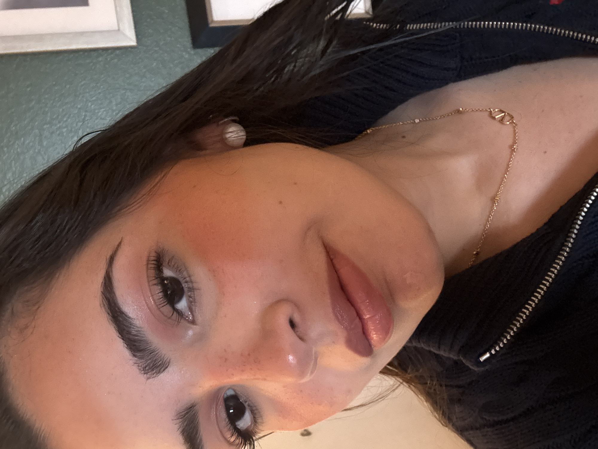 A close-up shot of a person with fair skin, dark hair, and defined eyebrows and eyelashes, likely indoors. The person is wearing a gold necklace with a distinctive 'V' logo and an earring. The skin appears to have makeup, including blush, and possibly lip gloss. The person is wearing a dark, possibly navy blue, zippered garment. The background is indistinct, with a hint of framed artwork on a wall in the upper portion of the image. There is no visible evidence indicating that the location is in San Antonio, United States of America.