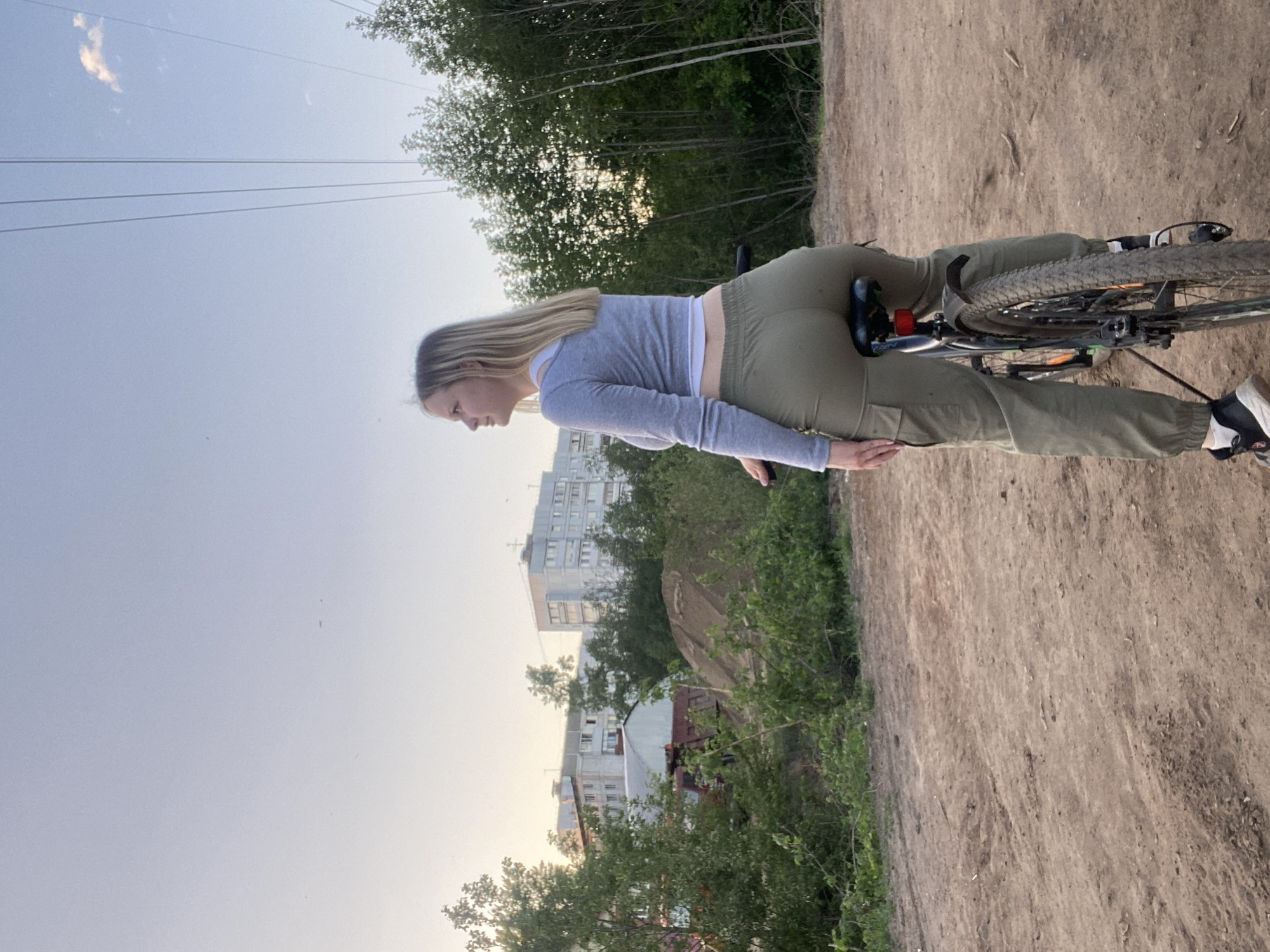 The image shows a young woman on a bicycle in Chaikovsky, Russia. She is wearing a grey long-sleeved crop top and olive green cargo pants. She has long blonde hair. The bike is a dark-colored mountain bike. The woman is standing still on a dirt path next to a green verge. In the background are buildings and trees. The sky is light blue with some wispy clouds. Electrical wires are visible in the upper left of the frame.