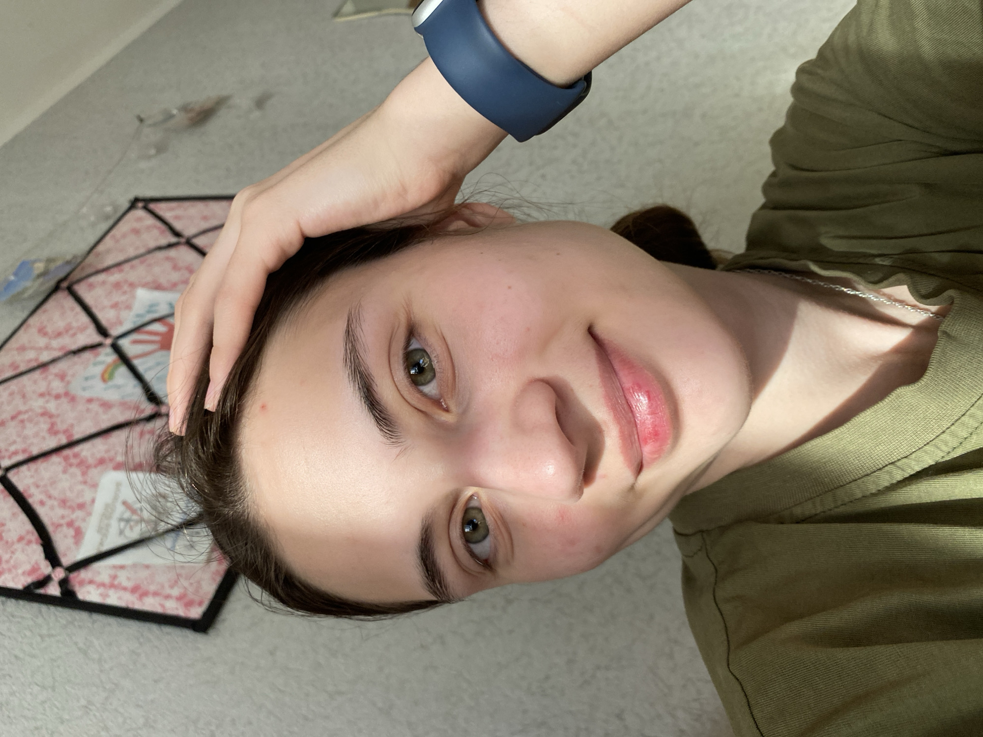 A person with fair skin, dark hair, and green eyes is looking at the camera with a slight smile. They have a blue smart watch on their left wrist, which is near their head. The person is wearing a green shirt. The background is a light-colored, patterned surface. A small, decorative object can be seen in the upper left corner of the image. The location is not specified and there is not enough information to determine if the scene is in the city of Votkinsk, Russia.