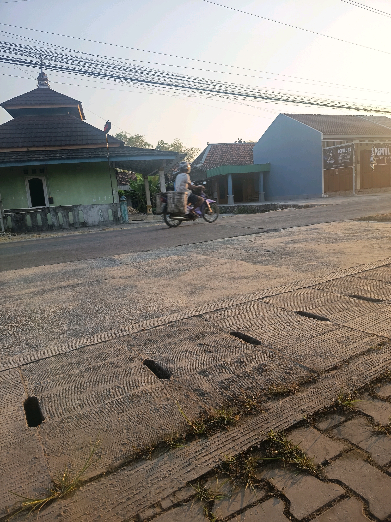 In Kalisari, Indonesia, a motorcyclist wearing a helmet and a white shirt rides a pink motorcycle on a paved road. The motorcycle is equipped with a large woven basket mounted on the rear. To the left of the road is a light green building with a dark brown roof, featuring a minaret, characteristic of a mosque. Adjacent to it, a darker blue building with a brown facade is visible, displaying signs advertising "RENTAL PS" and "RENTAL PS PARADISE," indicating a PlayStation rental business. Overhead, power lines crisscross the sky. The foreground of the image shows a textured concrete surface with rectangular openings, likely a drainage system, with patches of grass growing through the cracks. The scene is bathed in the warm, low light of either sunrise or sunset.
