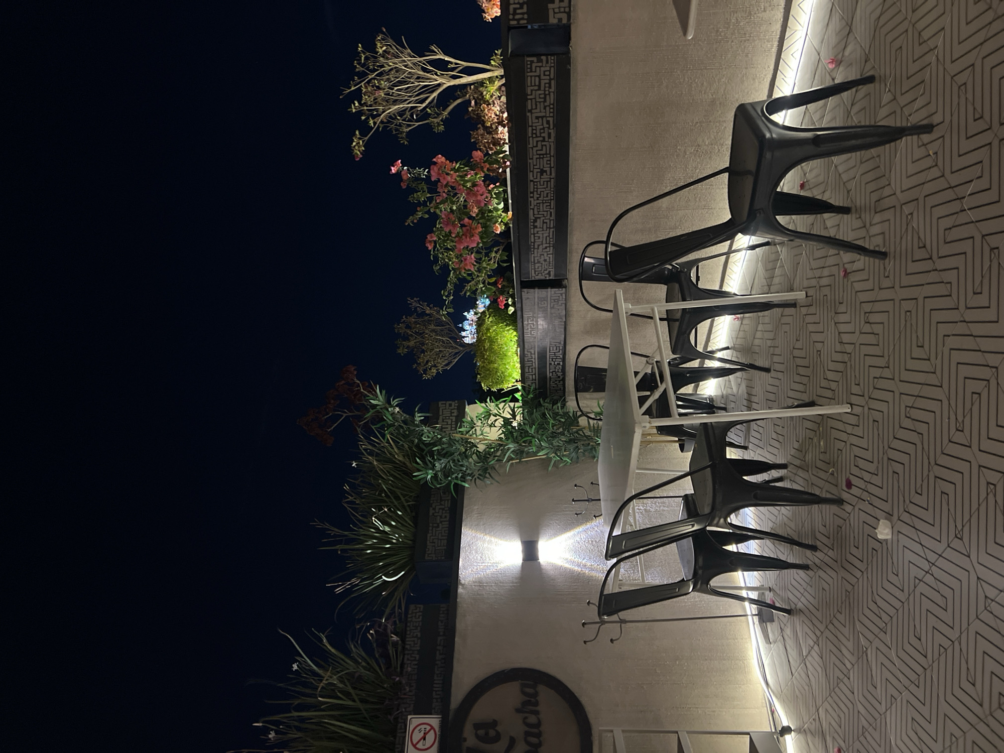 The image depicts an outdoor seating area, likely a patio, at a restaurant or cafe named "La Bacha". The location is in San Andrés Cholula, Mexico, based on the restaurant name. The time appears to be evening or night, indicated by the dark sky. Several empty black metal chairs are arranged on a patterned tile floor. One white folding table is present among the chairs. Plants and flowers are situated in planters along the wall. The planters are decorated with geometric patterns. A wall-mounted light fixture illuminates the area. A "no smoking" sign is visible near the bottom of the frame. No people are present in the image.