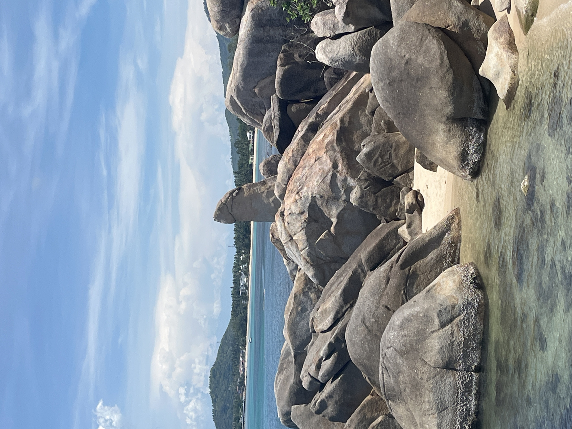 The image captures the Hin Ta and Hin Yai rocks, commonly referred to as Grandfather and Grandmother rocks, located in Baan Hua Thanon, Thailand. These distinctive rock formations sit beside a shoreline. The image is framed by the sky on the left and the ocean and the island shore in the background. The rocks themselves dominate the right side of the image. Water laps against the base of the rocks.