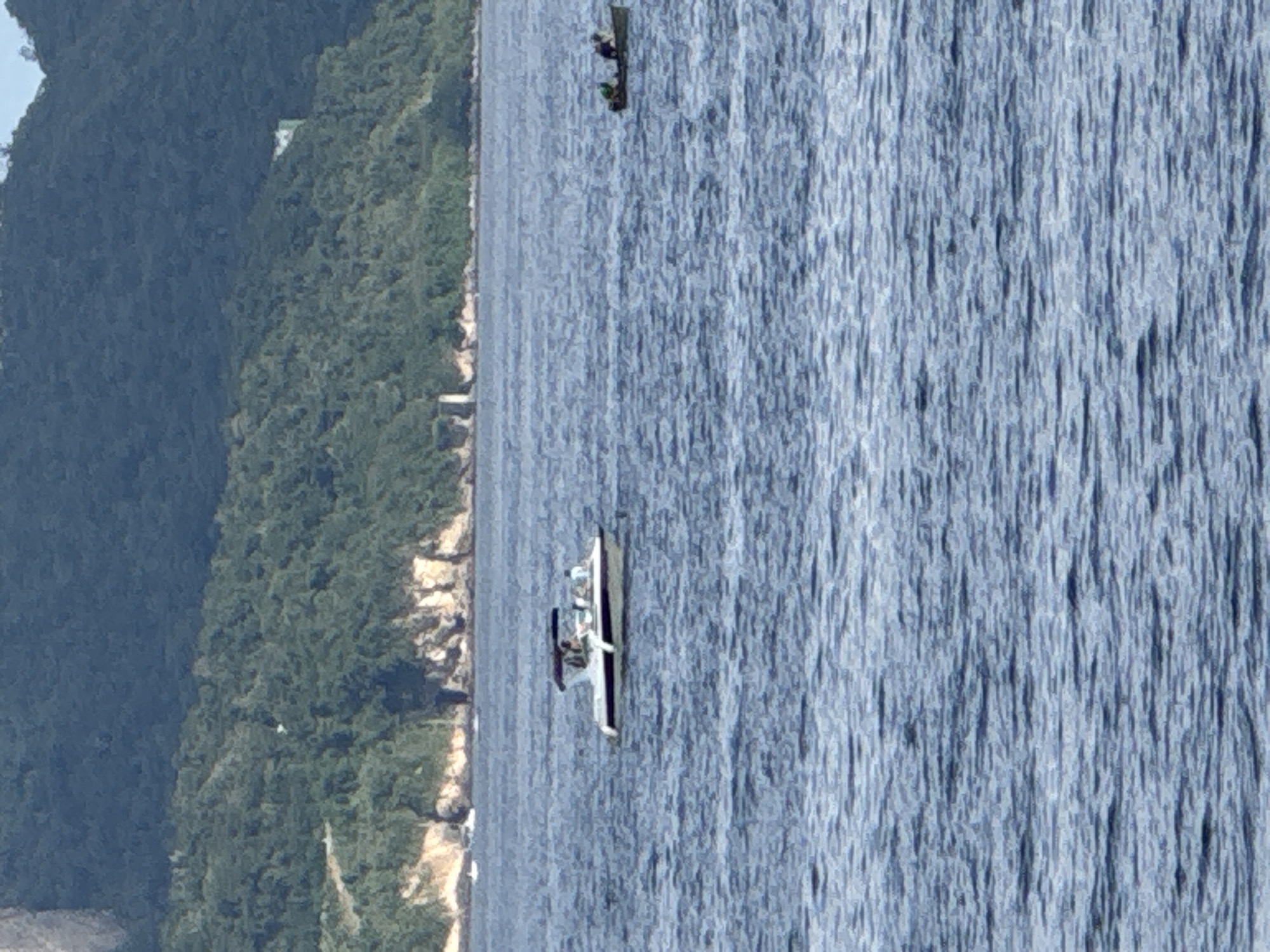 The image shows a wide view of the sea with a mountainous, densely vegetated coastline in the background. The sea is calm with ripples on the surface. Two boats are visible on the water.
In the mid-ground, a white motorboat with a canopy is positioned slightly to the left of center. The boat appears to have several occupants, though their features are indistinct. It is moving from left to right.
Further to the right and slightly closer to the background, a small, dark-colored boat, possibly a dinghy, is present. Two individuals are visible in this boat, one appearing to be operating it, while the other is seated.
The coastline behind the boats is characterized by steep, rocky cliffs with lush green vegetation covering the slopes. In the distance on the coastline, a few structures are discernible, including what appears to be a white building on the upper right side of the frame and some lighter-colored structures near the waterline on the left.
The overall impression is of a serene maritime scene with leisure activities taking place on the water against a natural, mountainous backdrop. This view is consistent with coastal areas of Niterói, Brazil, which features a prominent coastline with forested hills and mountains.