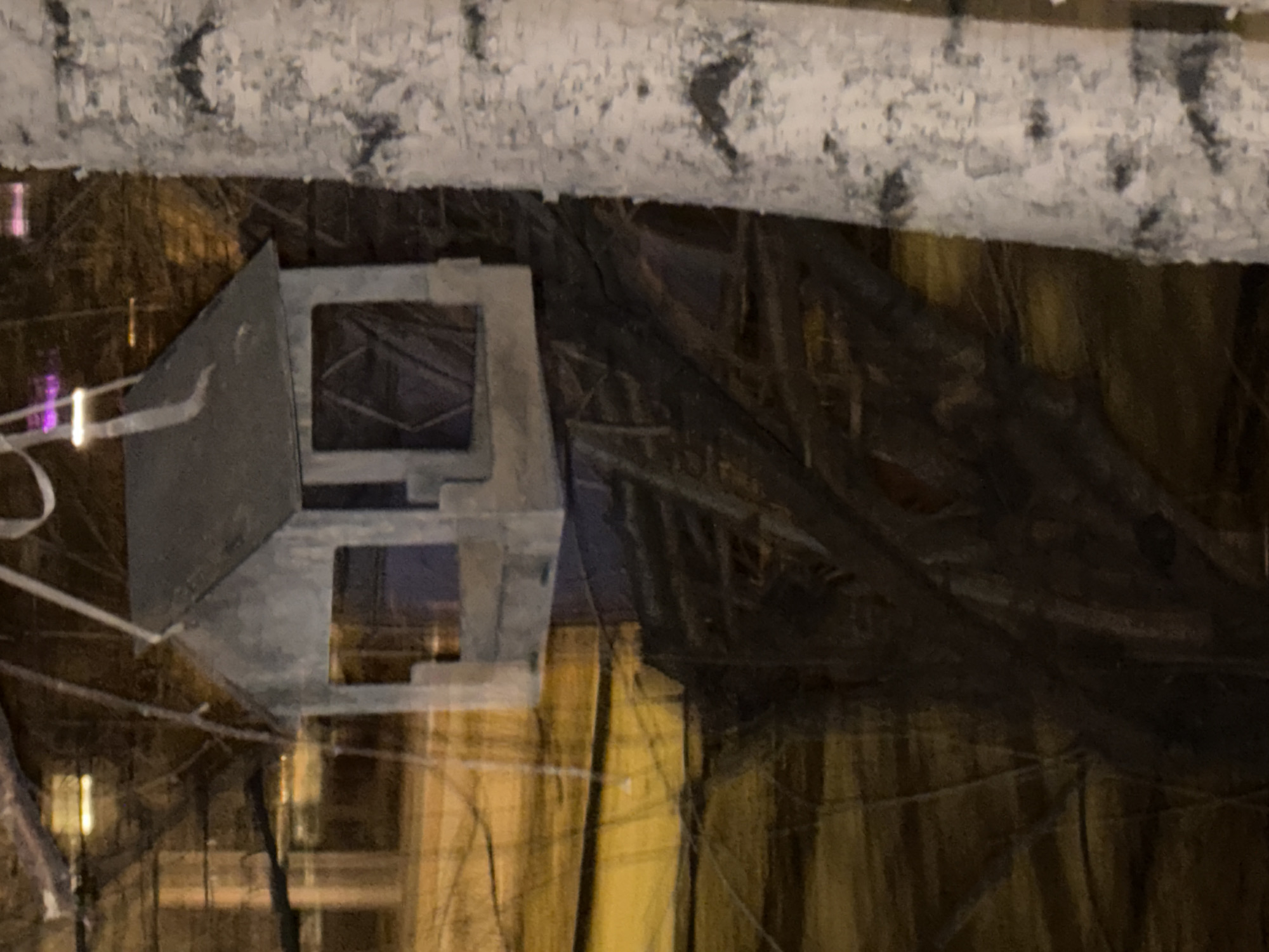 The image, taken in Krasnoyarsk, Russia, captures a close-up view of an artistic or decorative arrangement in what appears to be an indoor or dimly lit environment.
At the top of the frame, a horizontal log or beam, strongly resembling a birch trunk with its mottled white and grey bark, runs across the image. Its textured surface shows characteristic dark markings.
Below this log, slightly to the left of center, is a prominent light-grey geometric object. This object features a complex, angular design with multiple rectangular and square cut-outs, giving it a somewhat hollow or architectural appearance. It could be interpreted as a stylized building, an abstract house, or an intricate decorative element. Thin white lines or strings are visible attached to its left side, possibly used for suspension or as part of its construction.
The background is predominantly dark and indistinct, featuring textures that suggest draped fabric, netting, or a shadowed wall. Reflections of artificial light sources are visible within the darker areas, including a soft purple glow on the far left and warmer yellow/orange highlights scattered throughout, indicating an illuminated setting.
No people are present, and no specific activity is taking place. The scene is static, focusing on the interplay of shapes, textures, and subtle lighting. There is no visible text within the image.