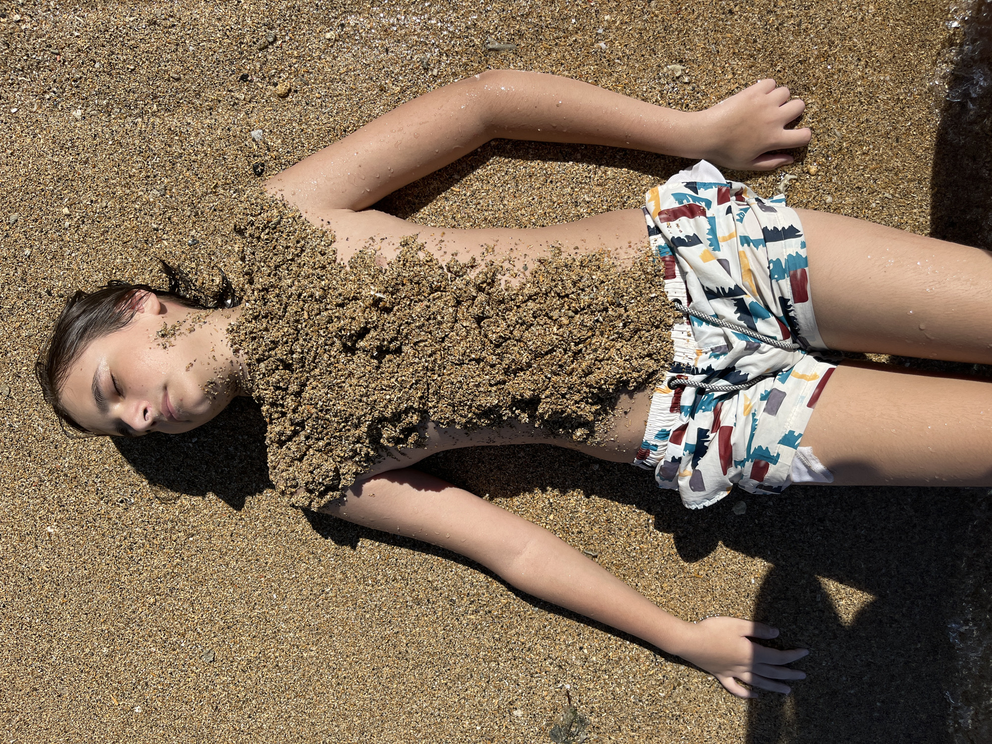 A young person is lying on a sandy beach, their body partially covered in sand. They are wearing patterned swim shorts. The person's eyes are closed and their head is tilted back, suggesting they are relaxed or sleeping. The beach appears to be near water, as indicated by the wet sand and a small wave lapping at the edge of the frame. The lighting suggests it is daytime, with the sun casting shadows on the sand. The scene is natural and appears to be a casual outdoor activity. The location is not specified in the image, and therefore cannot be identified as Denpasar, Indonesia.