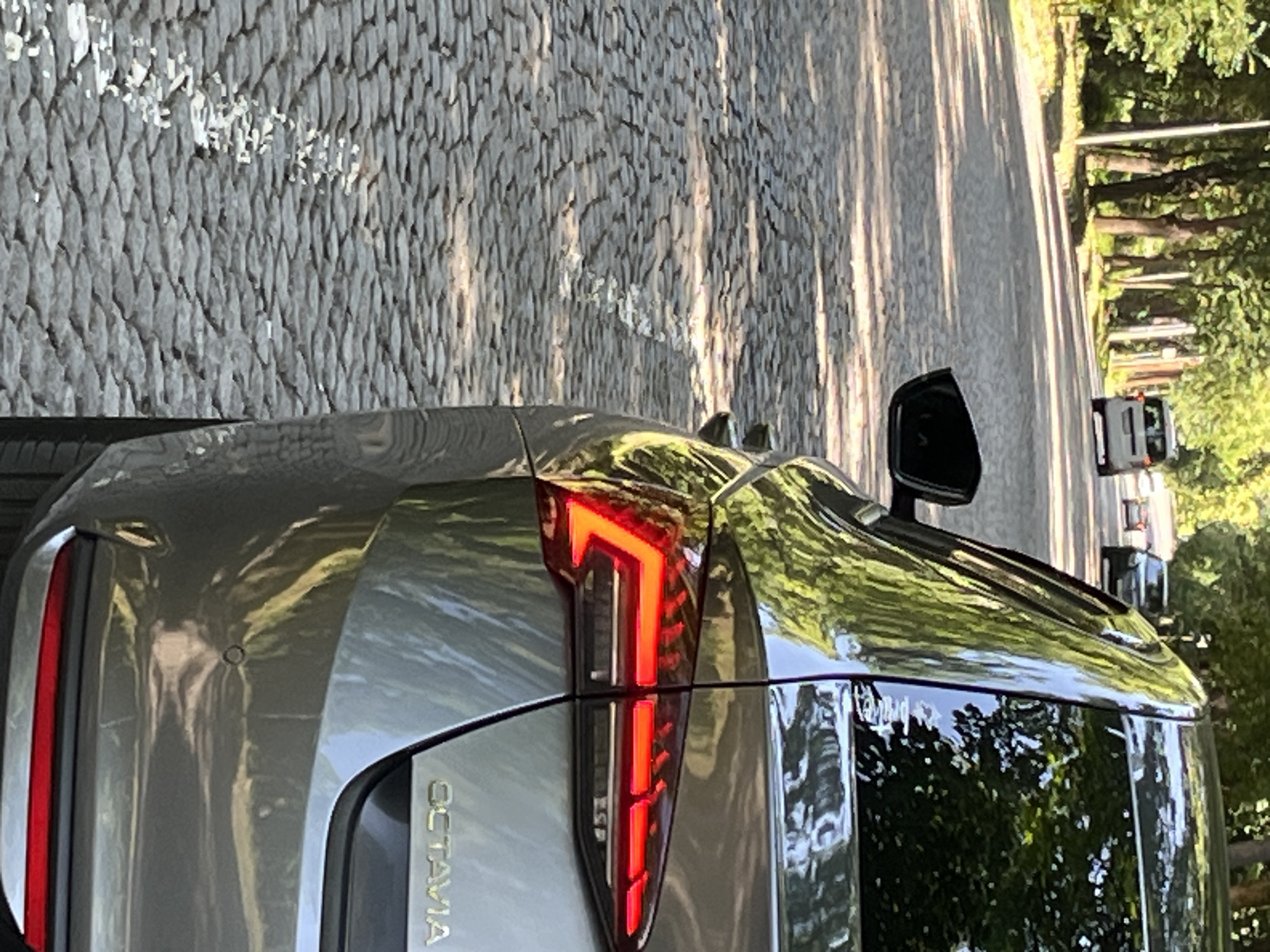 The image shows the rear of a silver Skoda Octavia parked on a cobblestone street in a park-like setting with trees lining the road. The car's taillights are illuminated, indicating it is either parked at night or its brake lights are activated. In the background, another white van and a dark-colored car are visible driving on the street. The cobblestone road suggests an older, possibly historic area of the city. The lighting indicates it is daytime, with sunlight casting shadows through the trees onto the road and car. The scene appears to be a peaceful, urban environment.