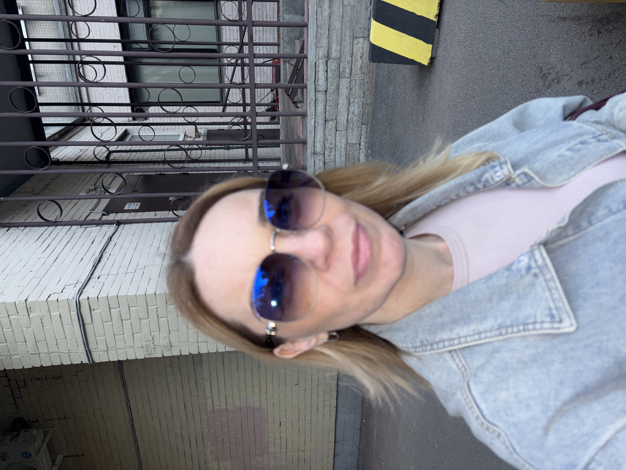 A woman is depicted taking a close-up selfie outdoors, with her head slightly tilted. She has blonde hair and is wearing large aviator-style sunglasses with reflective blue and purple lenses. Her expression is neutral, with her lips slightly parted. She is dressed in a light-colored top, possibly pink, and a light-wash denim jacket.
The background shows an urban setting. To the viewer's left (behind the woman's head), there is a light-colored brick building with multiple windows, some reflecting light. A dark metal security gate or grille with circular decorative elements is positioned in front of the building. To the right of the woman, a textured grey concrete wall or facade is visible, above a dark grey paved surface. A yellow and black striped curb or barrier is partially seen in the upper right portion of the background. Electrical wires run along both the brick and concrete walls. Faint, illegible markings are visible on the light brick wall to the right. An air conditioning unit can be seen on a lower part of the brick building in one of the provided crops.
The image appears to be taken during the daytime, possibly under an overcast sky or in a shaded area, given the ambient lighting and lack of harsh shadows. The activity is a selfie, with the woman looking directly towards the camera lens. The overall scene suggests an urban environment, possibly an alleyway or building exterior.