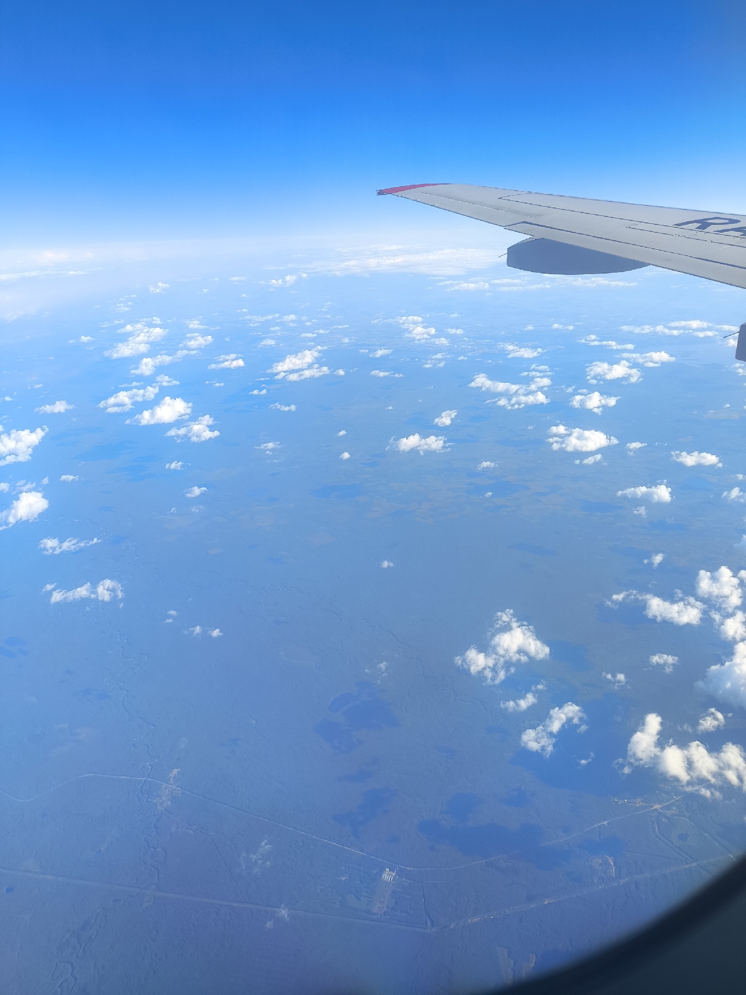 An aerial view from an aircraft window depicts a vast expanse of predominantly forested and wetland terrain under a clear blue sky, partially obscured by scattered cumulus clouds. The upper right quadrant of the image shows a section of a white airplane wing with a red tip on its leading edge, bearing partial text "R-" on its surface. Below the cloud layer, the landscape appears largely flat, characterized by extensive dark green forested areas and numerous irregular, dark blue bodies of water, indicative of lakes, ponds, and marshlands. Several linear features, resembling roads or cleared tracks, traverse the terrain. In the lower central portion of the image, a distinct cluster of light-colored structures and facilities is visible alongside one of these linear features, suggesting a small industrial site or settlement. This scene is observed over the region of Khimki, Russia, showcasing the broad natural landscape surrounding the city, including its characteristic forests and wetlands, with minimal visible urban development from this altitude. No people or specific actions are discernible.