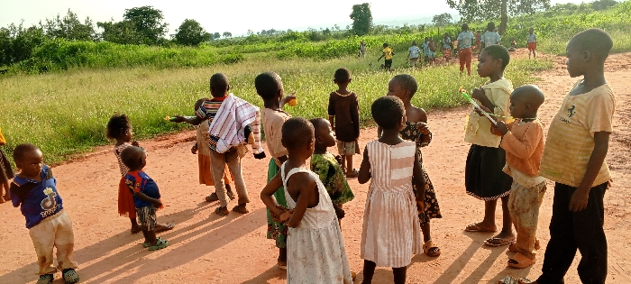 An outdoor scene in Ngalula, Democratic Republic of the Congo, depicts approximately 18-20 children on and around a reddish-brown dirt path. In the foreground, a group of about 15 children, varying in age from toddlers to early teenagers, are gathered on the path. Their skin tone is dark, and they are dressed in casual, light-colored clothing, including t-shirts, shorts, and dresses.
Specific individuals observed include:
* A young boy on the far left wearing a blue t-shirt with a yellow emblem and light-colored trousers; his lower arms are not visible.
* A smaller child next to him wears a blue top and red shorts.
* A child in the mid-left holds a yellow, spherical object.
* Two children on the mid-right are holding slender, colorful objects, possibly toys such as bubble wands. One wears a light yellow short-sleeved top and a dark skirt, while the other wears an orange long-sleeved shirt and light-colored trousers.
* A girl in a light-colored, vertically striped dress stands towards the right.
* A taller boy on the far right wears a striped t-shirt with a yellow logo and dark trousers.
Most children in the foreground are observing their surroundings or subtly interacting within the group. Some are barefoot, while others wear sandals or crocs.
The environment consists of the dirt path, which shows tire tracks or depressions. Lush green grass and denser vegetation, including bushes and trees, border the path and extend into the midground. In the background, more children are visible within a field of green vegetation, some appearing to be running or playing. The horizon reveals distant rolling hills under a bright, clear sky. Shadows are visible on the path, indicating daylight.
