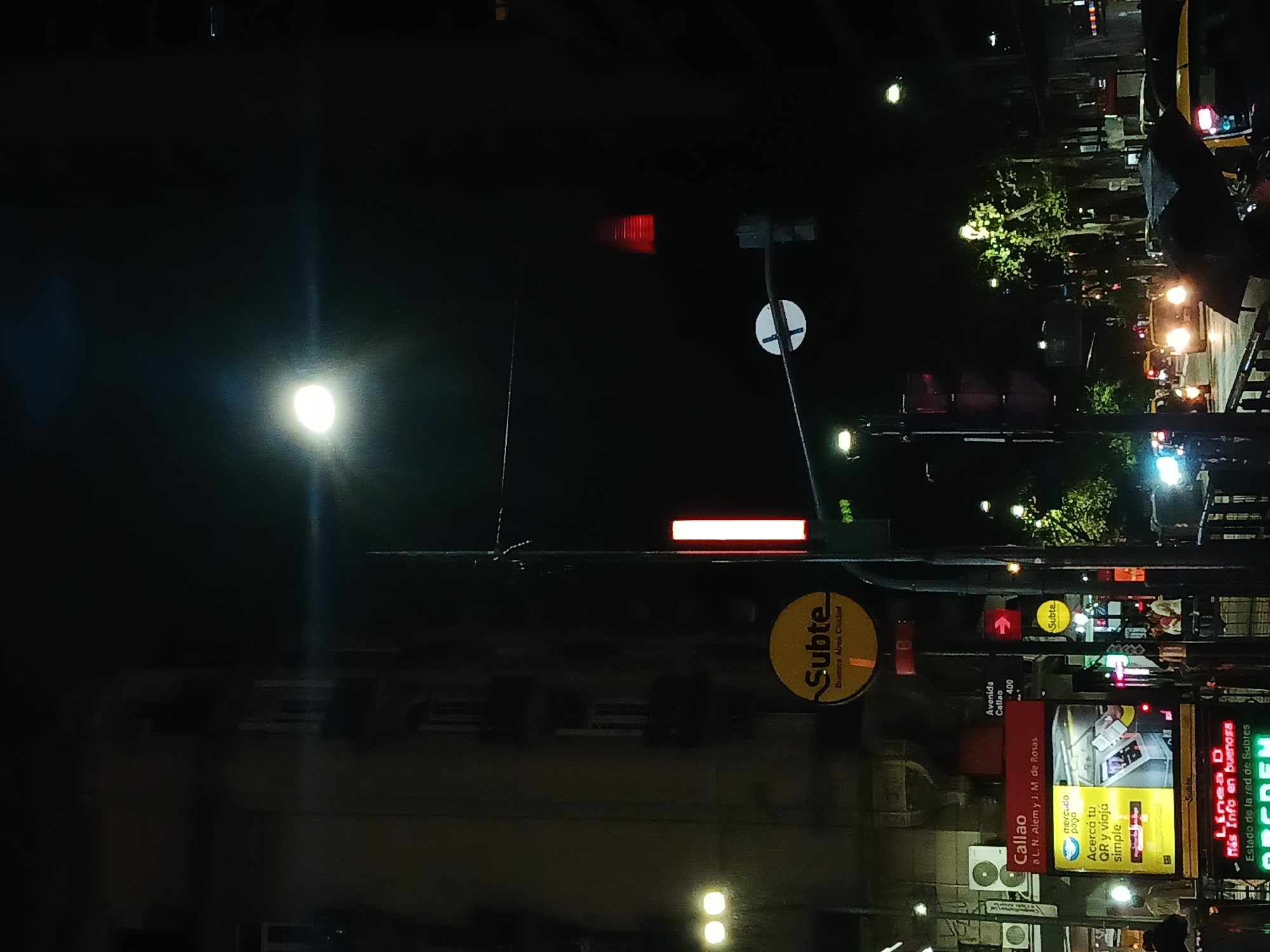 The image depicts a street scene at night in Buenos Aires, Argentina. Streetlights illuminate the scene. A prominent sign advertises the Subte (subway), indicating the entrance to the Callao station. The advertisement includes a QR code for easy payment and travel. Another sign shows the location of the Line D of the Subte, providing information on its status. The intersection of Avenida Callao with Leandro N. Alem and Juan Manuel de Rosas is nearby. The surrounding buildings and street furniture are silhouetted against the night sky. The scene suggests a busy urban environment with active public transportation.