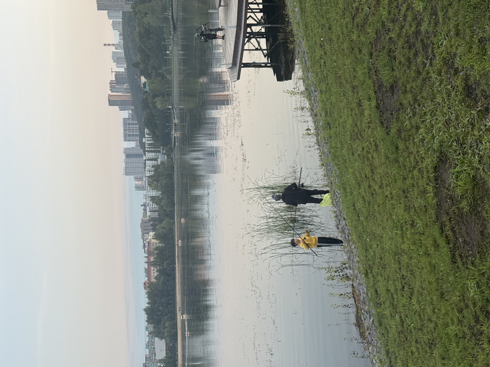 The image captures a serene waterscape with a cityscape in the background. The scene is dominated by a calm body of water, reflecting the overcast sky and the distant urban skyline. On the right side of the frame, a grassy embankment slopes down towards the water's edge, with a path or small clearing visible.
In the foreground, two individuals are engaged in fishing near the reeds. One person, wearing a yellow jacket, stands closer to the camera, holding a fishing rod. Another person, dressed in darker clothing, stands further back, also fishing. Their postures suggest concentration and quiet pursuit.
The background reveals a dense collection of tall buildings, indicative of an urban environment. Trees line the waterfront between the city and the water. The sky is a uniform light grey, suggesting either early morning or late evening, or a day with significant cloud cover. The overall mood is tranquil and peaceful, with the natural elements of water and grass juxtaposed against the man-made structures of the city. No visible text is present in the image. The location is identified as Ufa, Russia.