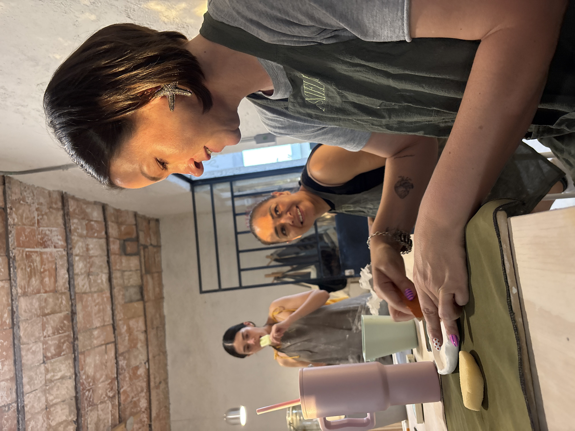 The image shows three women engaged in what appears to be a pottery or ceramics class or workshop. The woman in the foreground is leaning over a table, her hands manipulating a tool and some clay-like material. She has short, dark hair with a star-shaped earring and is wearing a gray t-shirt with a dark green apron. The table surface is covered with a light green material, and there are ceramic mugs and other tools present.
Behind her, another woman is visible, smiling and looking towards the camera. She is wearing a dark tank top. Further back, another woman is working at a table with a light-colored apron.
The location appears to be inside a building, possibly a studio or workshop. A section of exposed brick wall is visible on the left side of the frame. A window with dark frames can be seen in the background.
The image was taken in San Bernardino Tlaxcalancingo, Mexico.