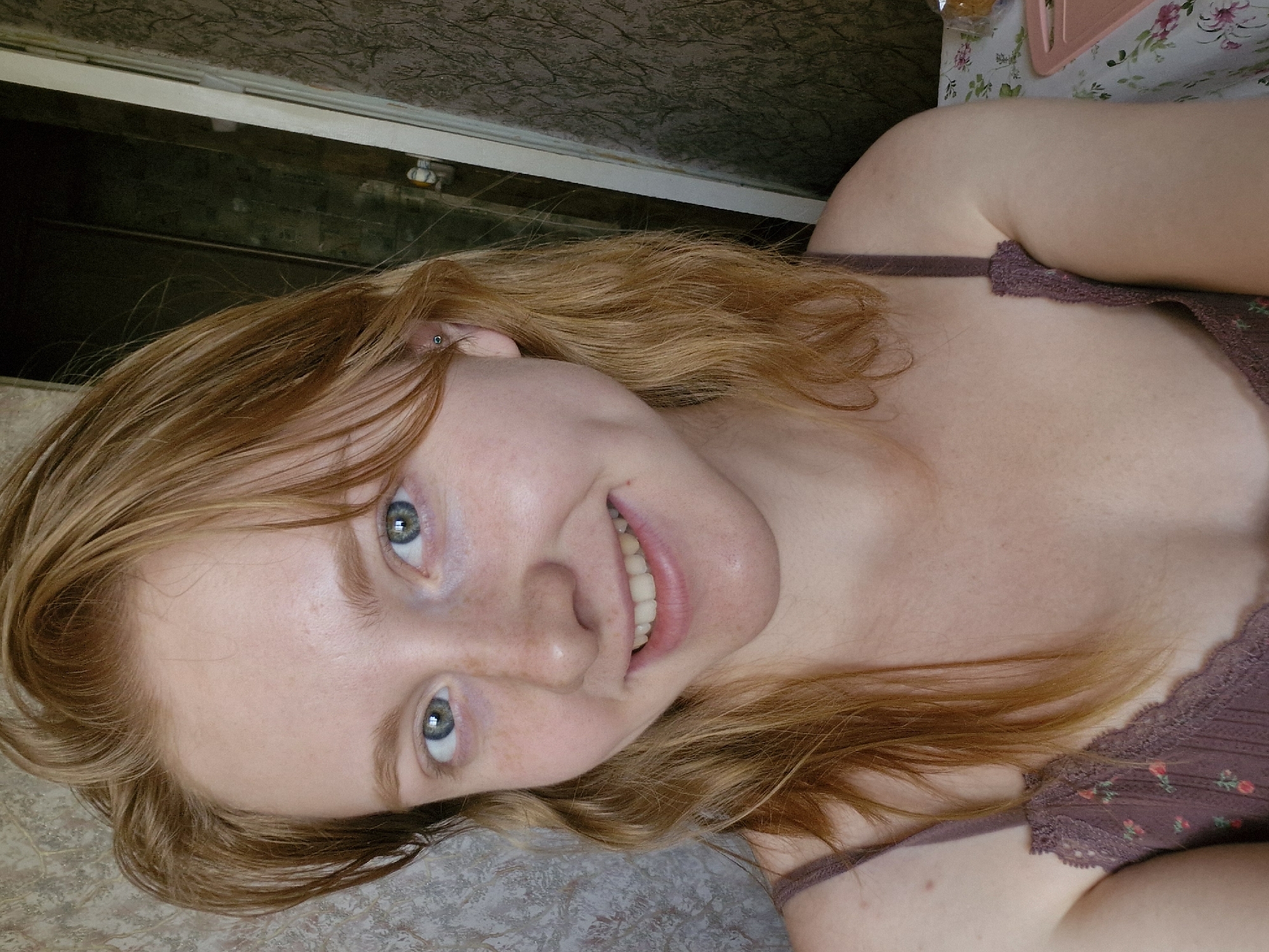 A woman with strawberry-blonde hair and blue eyes lies down, smiling at the camera. She has freckles on her face. She wears a brown floral-patterned top with lace trim. An earring is visible in her left ear. A patterned surface is underneath her. A door and a floral-patterned object are partially visible in the background. There is no evidence to suggest that this image was taken in Yekaterinburg, Russia.