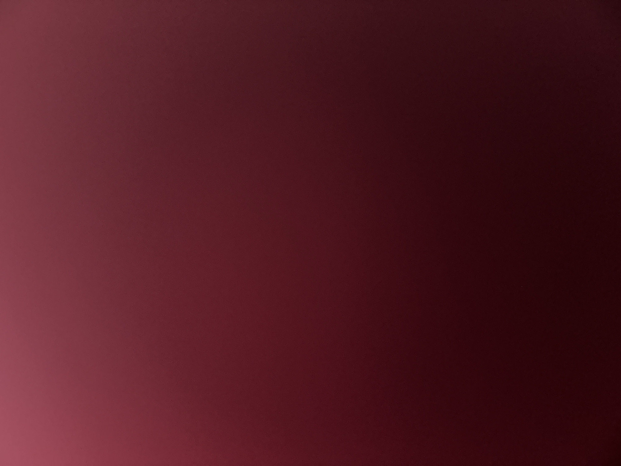 This is a medium shot, horizontal, full-frame image of a textured, dark reddish-brown surface. The lighting creates a gradient effect, with the bottom left corner being the brightest, a lighter shade of red, and the color gradually darkening towards the top and right edges, becoming a deep, almost black, brown. There are no discernible subjects, objects, or scenes. The image appears to be an abstract representation of a color or texture, possibly a close-up of fabric, a wall, or a painted surface. There is no visible text, time of day, weather, or location cues. The overall mood is subdued and perhaps a bit moody due to the deep color saturation.