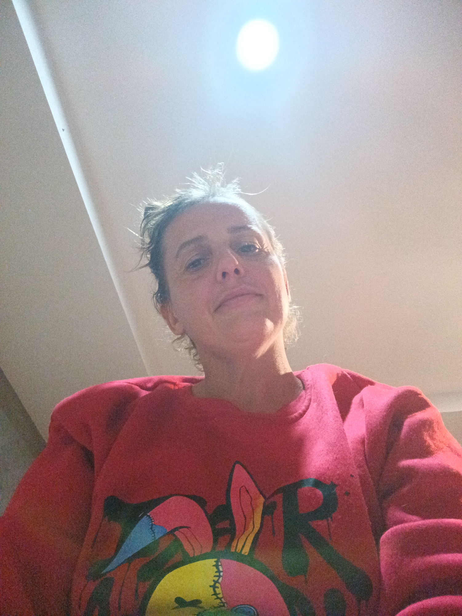 A woman is looking down at the camera. She has light skin and her hair is pulled back. She is wearing a red sweatshirt with a colorful graphic on the front. The background is a white ceiling with a light fixture. The location in the city of Ban Tak Daet, Thailand is unknown.
