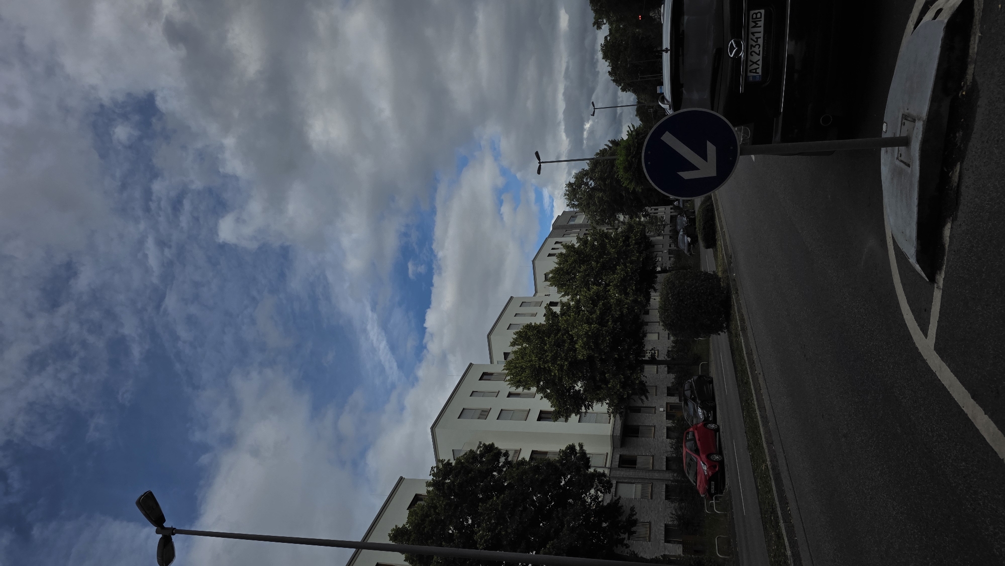 The image depicts a street scene in a city. On the left side of the street, a row of modern white apartment buildings with balconies lines the sidewalk, interspersed with mature trees. A street lamp stands tall on the left. A red car and a black car are parked on the side of the road. On the right side of the street, a black SUV with a license plate reading "AX 2341 MB" is parked. A blue circular traffic sign with a white arrow pointing left is prominently displayed in the foreground. The sky above is a mix of blue patches and overcast clouds. The street is asphalt, with white markings indicating lanes and a roundabout. The overall impression is one of a residential urban area.