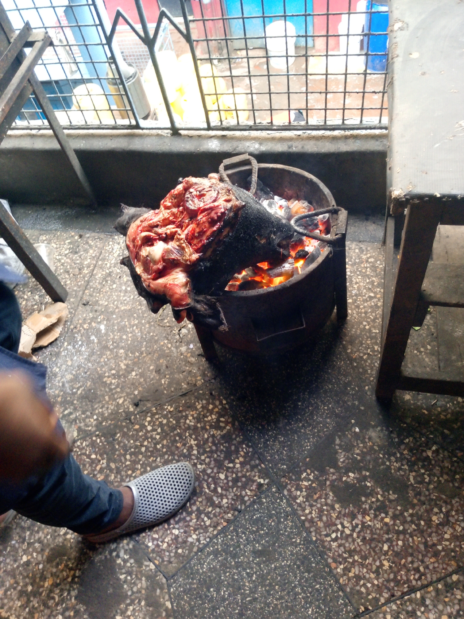 The image shows what appears to be a goat head being roasted over an open charcoal fire in an urban setting. The head is partially charred, with visible meat and skin. The fire is contained in a metal stove. A person wearing jeans and gray perforated shoes stands nearby. The floor is tiled with a speckled pattern. In the background, there is a window with a metal security grill. The location is indicated by the environment to be Nairobi, Kenya.