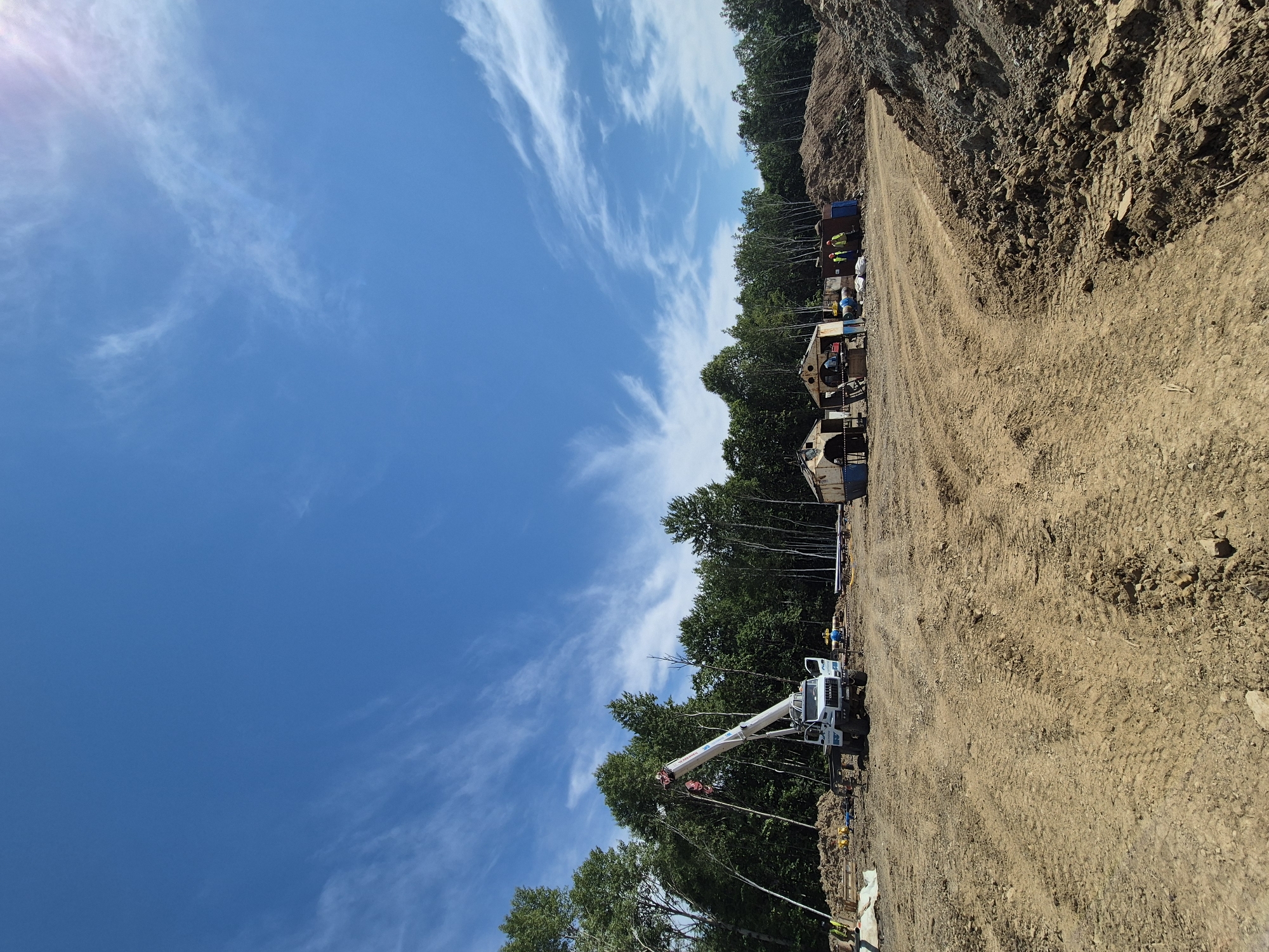 A construction site is depicted under a bright blue sky with wispy clouds. The foreground is dominated by a dirt track with tire marks. In the mid-ground, a white crane truck is parked on the muddy ground. To the right of the crane, several portable structures or sheds are visible, some with open doorways. A pile of reddish-brown earth is located to the right of these structures. In the background, a dense forest of green trees lines the horizon. Several individuals in yellow vests are present on the site, engaged in work or observation. The scene appears to be a construction project in progress in a rural or undeveloped area, possibly near Taezhnoe, Russia, given the mention of the city.