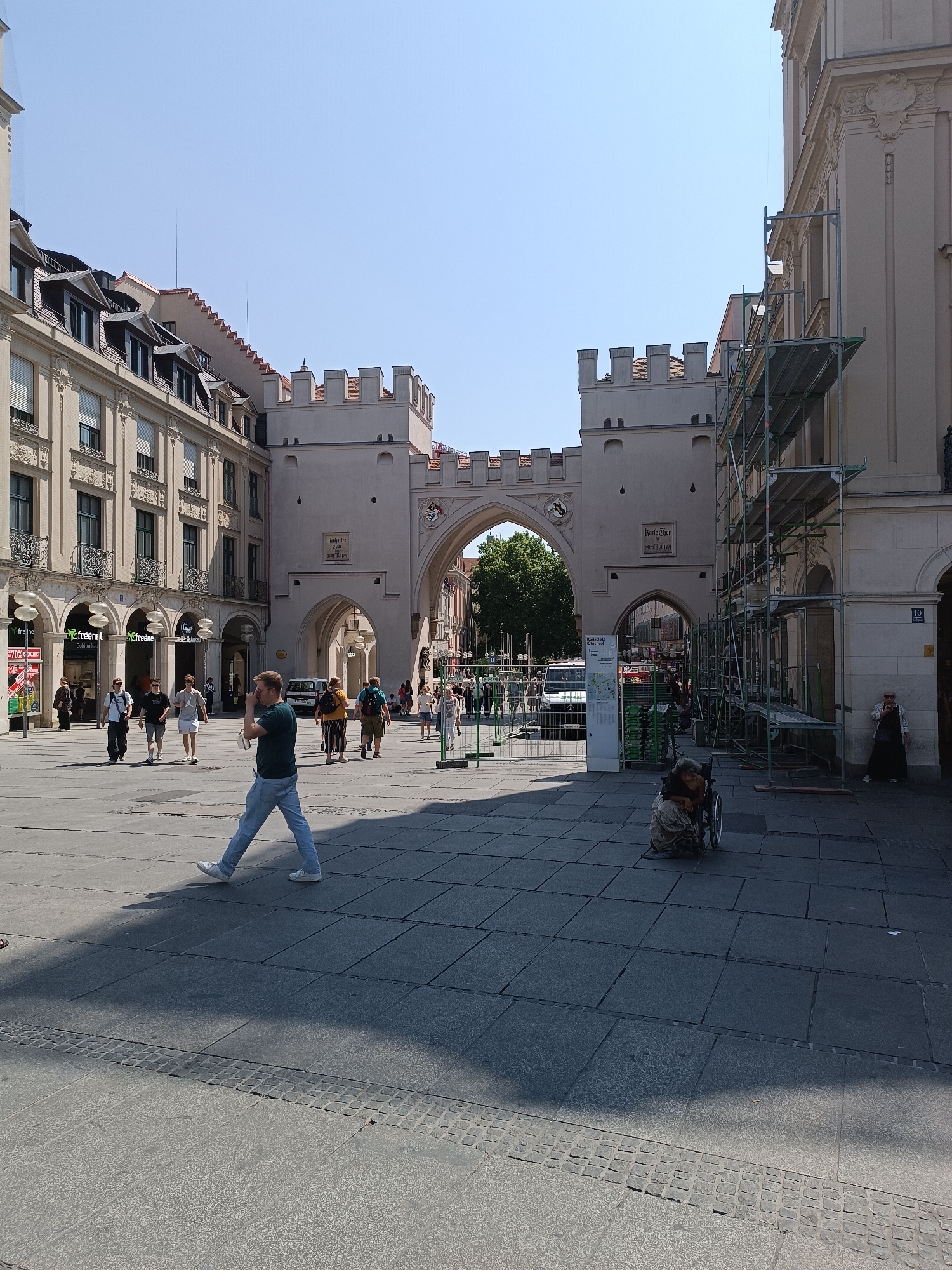 The image shows a wide, paved city square with a large, medieval-style stone archway dominating the background. The archway has crenellated battlements and appears to be part of a historical building. Scaffolding is erected on the right side of the archway, suggesting ongoing construction or renovation.
In the foreground, several people are visible. On the left, a man in a green shirt and blue jeans walks towards the viewer, his head turned away. Further back, a group of young people walks together. To the right of the archway, in the middle ground, a person in a wheelchair is seated on the ground, seemingly interacting with someone also in the wheelchair or leaning over them. Another woman in a black skirt and white top stands to the far right, near the scaffolding.
The square is paved with large, grey stone slabs, and there are some smaller cobblestones forming a border in the foreground. The sky is clear and blue, indicating a sunny day. Shadows are cast across the square, indicating the time of day.
This scene is likely located in Munich, Germany, at the Sendlinger Tor, a historical city gate that is a prominent landmark and public square in the city. The architecture of the archway and the surrounding buildings are consistent with the style found in Munich's historical center.