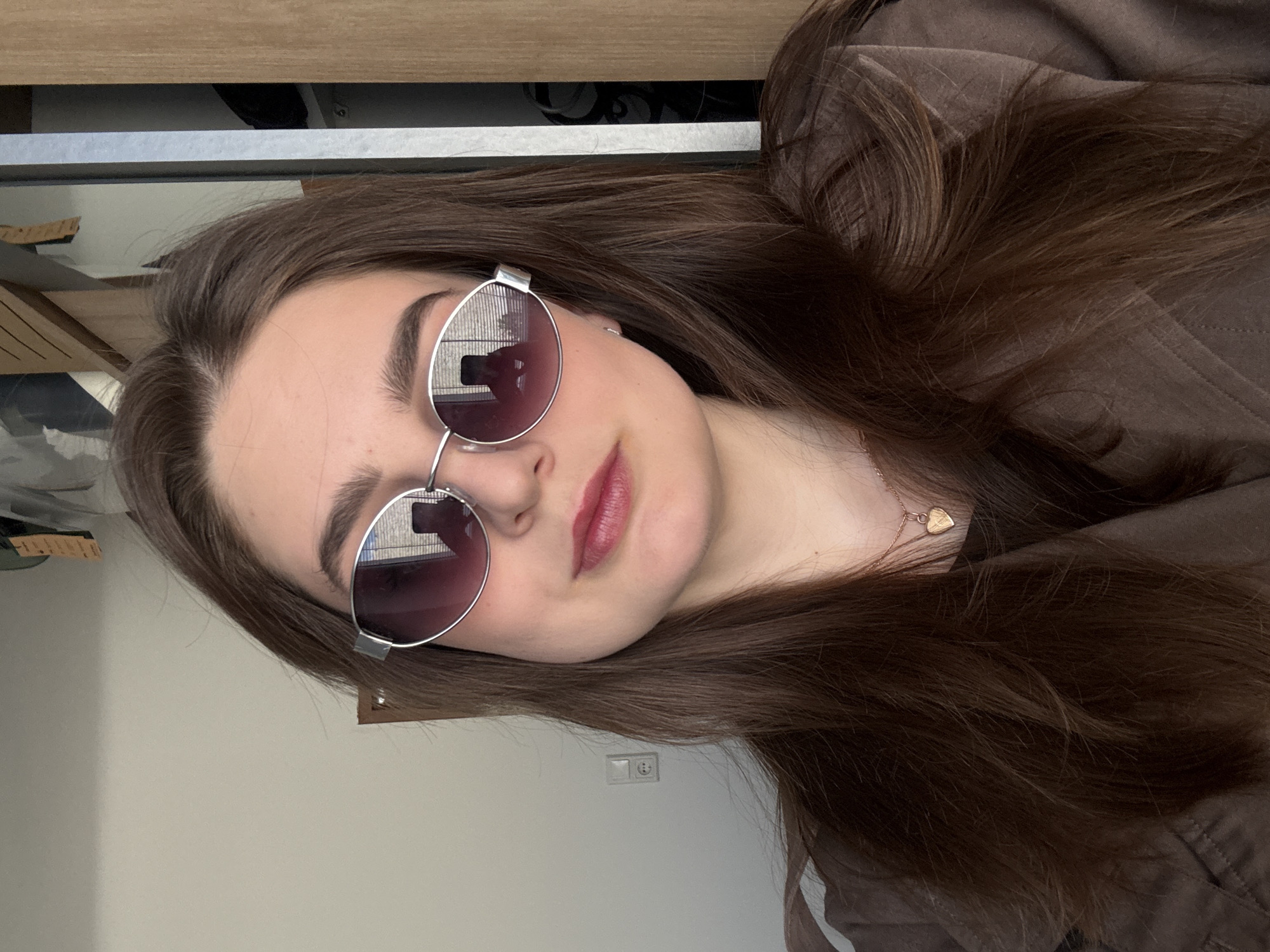 A close-up portrait shows a young individual, likely female, with long, straight, dark brown hair, reclining indoors. The subject wears round, silver-framed sunglasses with dark, reflective lenses, obscuring her eyes. Her lips are made up with a reddish-pink lipstick, and her eyebrows are dark and well-defined. She is wearing a dark brown, possibly quilted, jacket or hoodie, and a delicate gold-colored necklace with a small heart-shaped pendant is visible around her neck.
The background consists of a light-colored wall with a white electrical outlet visible, and a wooden surface along with a grey horizontal bar, suggesting interior furniture or architectural details. Reflections in the sunglasses reveal distorted views of the indoor environment, including what appears to be structural lines or window frames. The lighting is soft and even, indicative of an indoor setting during the daytime. The overall mood is relaxed and casual. There is no visible text in the image. The location is Hainburg an der Donau, Austria.
