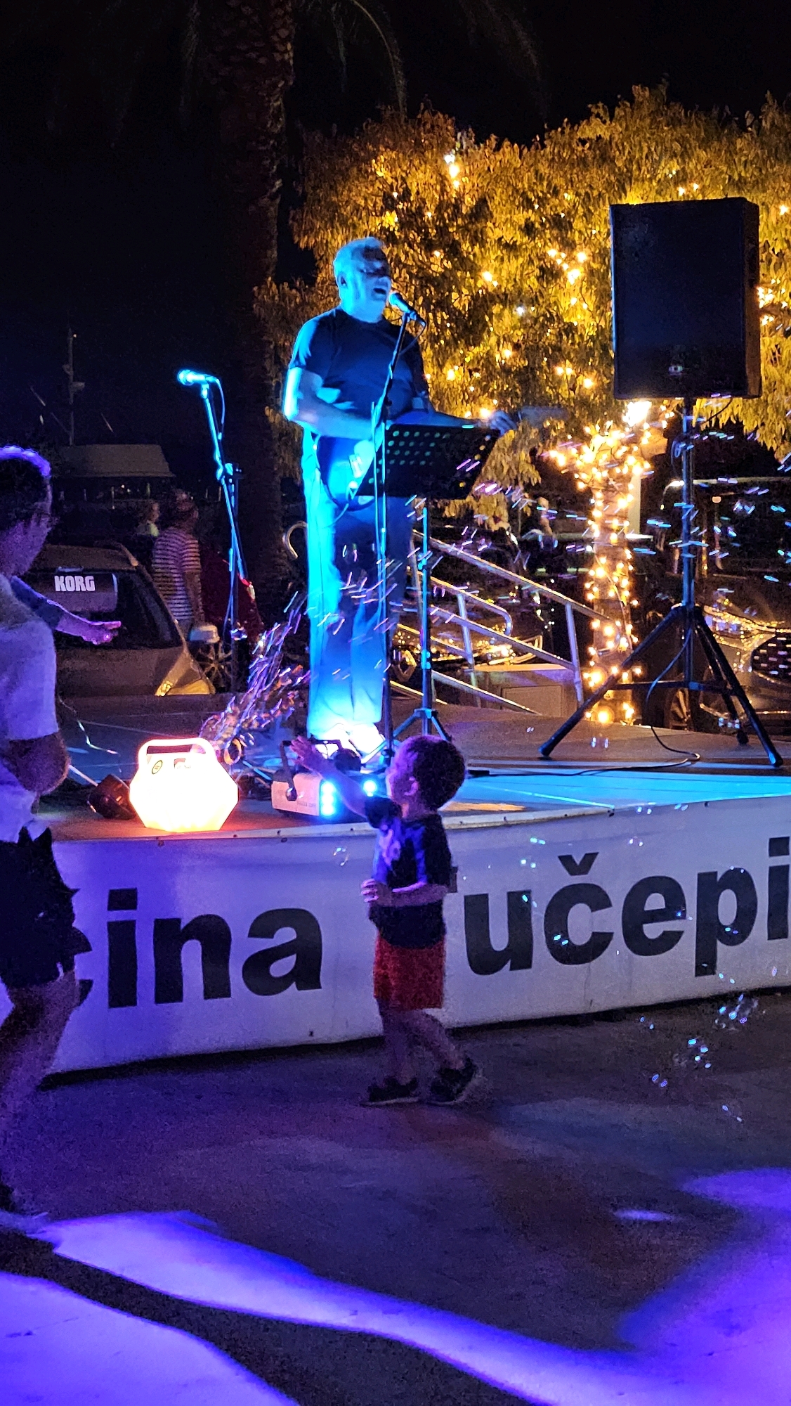 A full shot captures an outdoor nighttime event in Općina Tučepi, Croatia, where a man is performing on a stage illuminated by blue and yellow lights. The man, wearing a black t-shirt and jeans, is playing a guitar and singing into a microphone. He stands behind a music stand, with a speaker to his right and a palm tree and a tree decorated with numerous yellow lights behind him.
In the foreground, a young boy in a black t-shirt and red shorts reaches out towards a stream of bubbles, his face filled with wonder. To his left, a hexagonal, glowing white lantern sits on the stage. The stage itself has a banner with dark lettering that appears to read "cina učepi," likely part of the location's name. A person with their back to the camera, wearing a white t-shirt, is visible to the far left, also seemingly interacting with the bubbles. In the background, a vehicle with the "KORG" logo is parked, and other people can be seen in the distance, suggesting a lively gathering. The ground is dark asphalt, reflecting the purple and blue stage lighting.