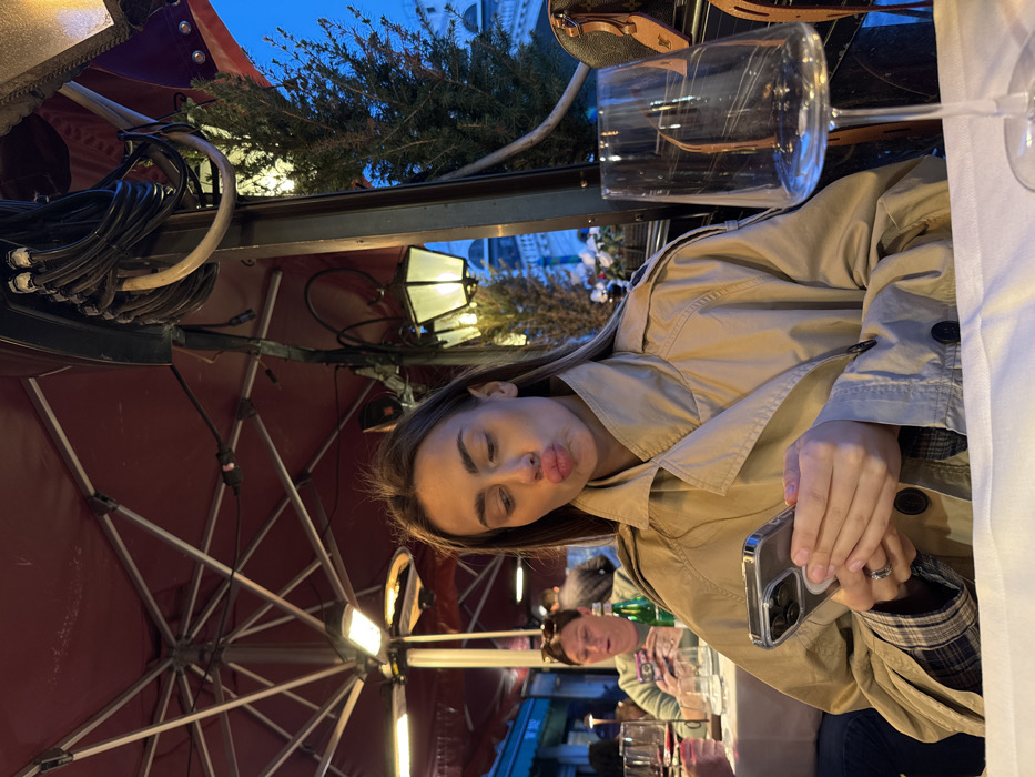 A young adult female is depicted seated at an outdoor dining table, likely a restaurant or cafe in Venice, Italy, during twilight. She wears a light brown trench coat over a plaid shirt and holds a silver-cased smartphone with both hands. Her lips are pursed in a "duck face" expression, directed towards the viewer. A ring is visible on her left ring finger. The table is covered with a white tablecloth, and an empty stemmed wine glass is prominently positioned in the right foreground. Overhead, a large reddish-brown canopy or umbrella provides shelter, fitted with a radiant heater. To the right, a dark structural pole supports a decorative street lamp and features bundled electrical cables. Adjacent to the pole is a potted evergreen plant. In the mid-ground, another female is seated at a separate table, focused on her own smartphone. A green bottle is present on her table. The background displays blurred architectural details, including a dome-like structure, beneath a deep blue sky. A brown designer handbag is visible on the far right.