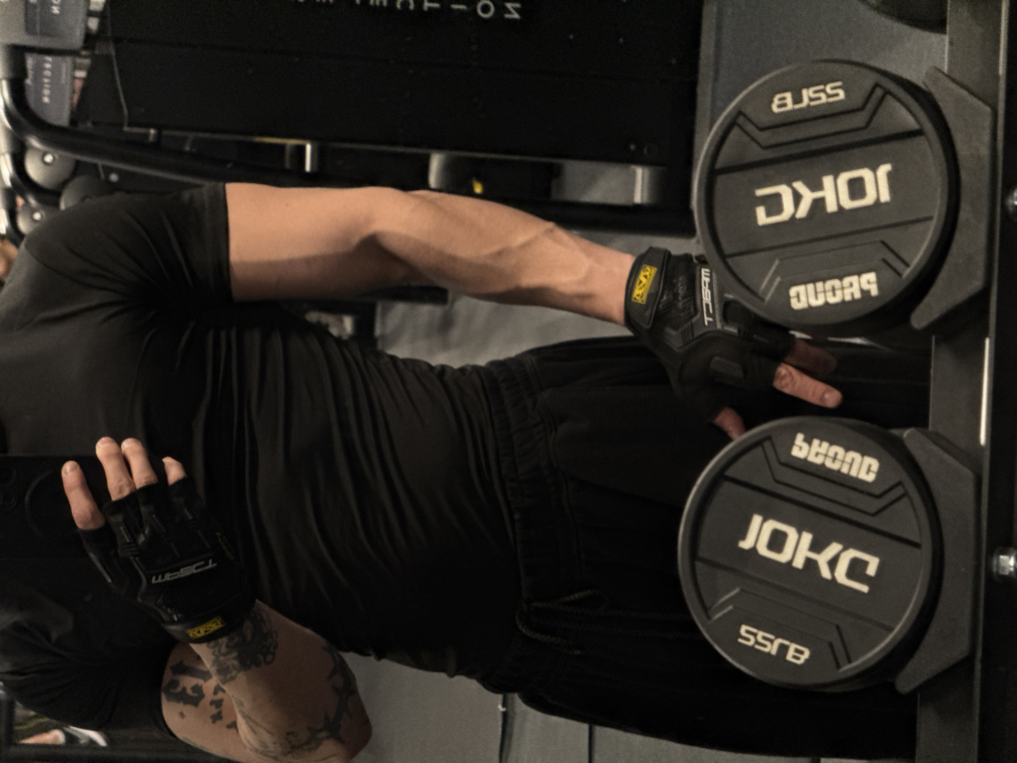 A man in a black outfit, including workout gloves, is standing in what appears to be a gym. He is facing a weight rack that holds JOKC-branded weights, with "SSRB" and "BEYOND" inscribed on them. The environment is dimly lit, and the scene is tightly framed. The man's arms display prominent veins, suggesting physical exertion. No specific location in Sofia, Bulgaria, is discernible from the image.