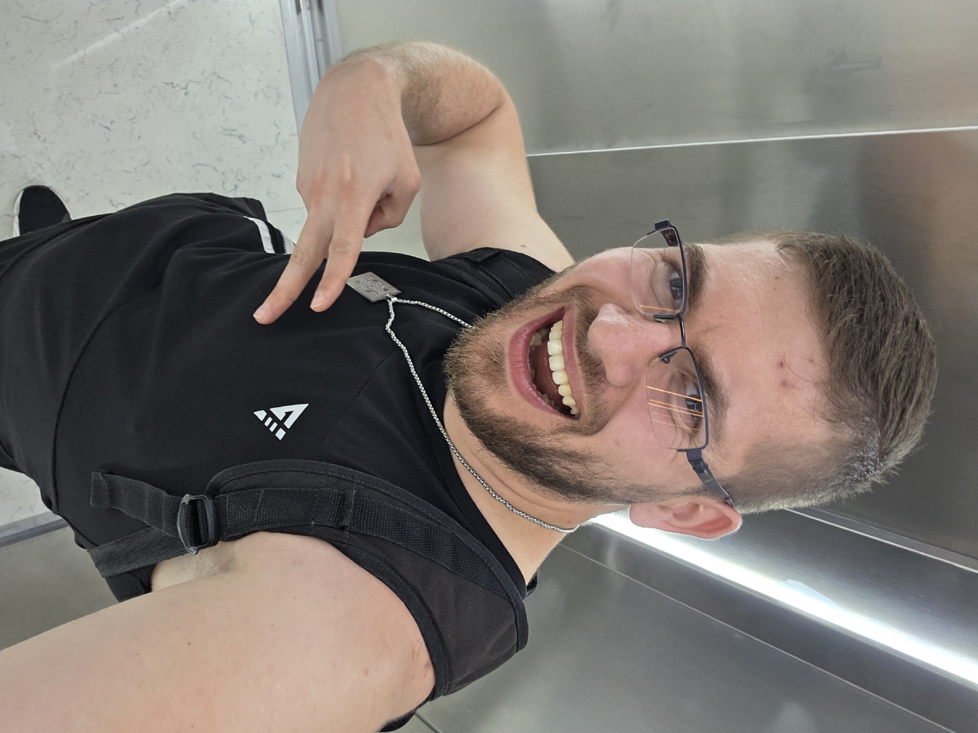 A man stands in what appears to be an elevator, indicated by the stainless steel walls and lighting. The floor has a marble pattern. He is wearing a black t-shirt, black pants with white stripes, and black shoes. A black strap is slung over his shoulder, possibly part of a bag or backpack. He wears glasses and a silver chain necklace. He is making a hand gesture with his index and middle fingers extended. His facial expression shows a wide grin. The location is not discernible from the image.
