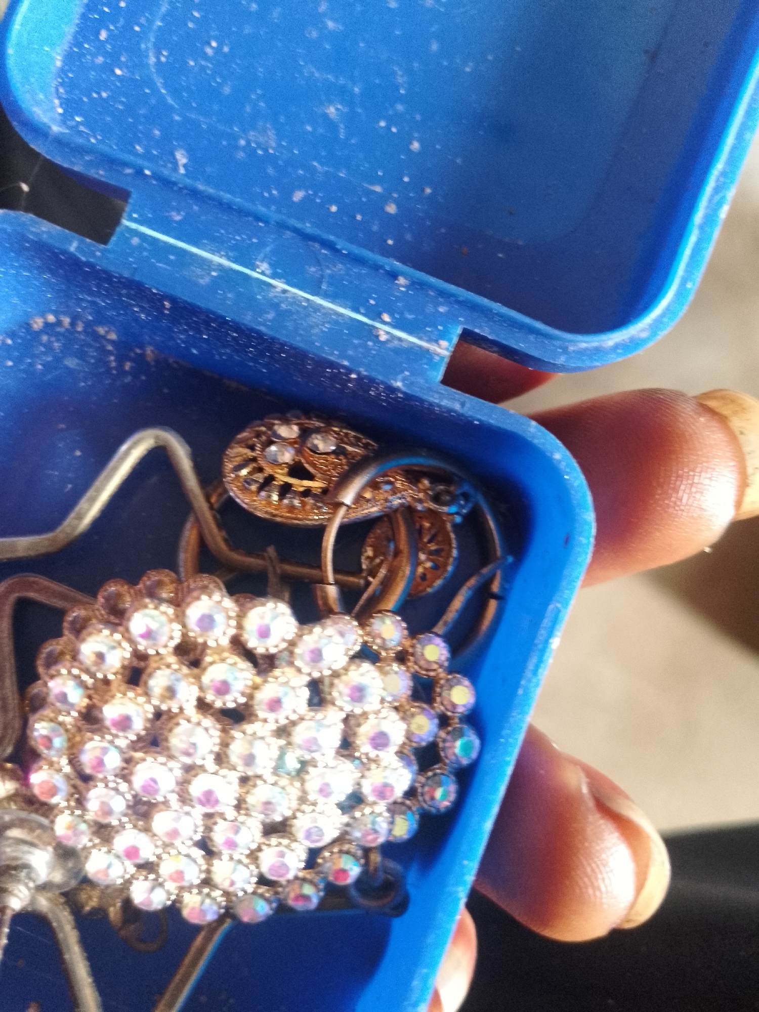 A close-up, overhead shot shows a blue plastic container filled with jewelry. The container appears dusty. In the foreground, a large brooch or pendant with numerous clear, iridescent rhinestones is visible. Behind it, a smaller, gold-colored circular piece of jewelry with inset stones can be seen, along with other metallic elements of jewelry, possibly rings or chains. A person's thumb and forefinger, with trimmed fingernails, are holding the container from the right side. The background is blurred, showing a light-colored surface. The image is a detailed, forensic, matter-of-fact depiction of a collection of jewelry.