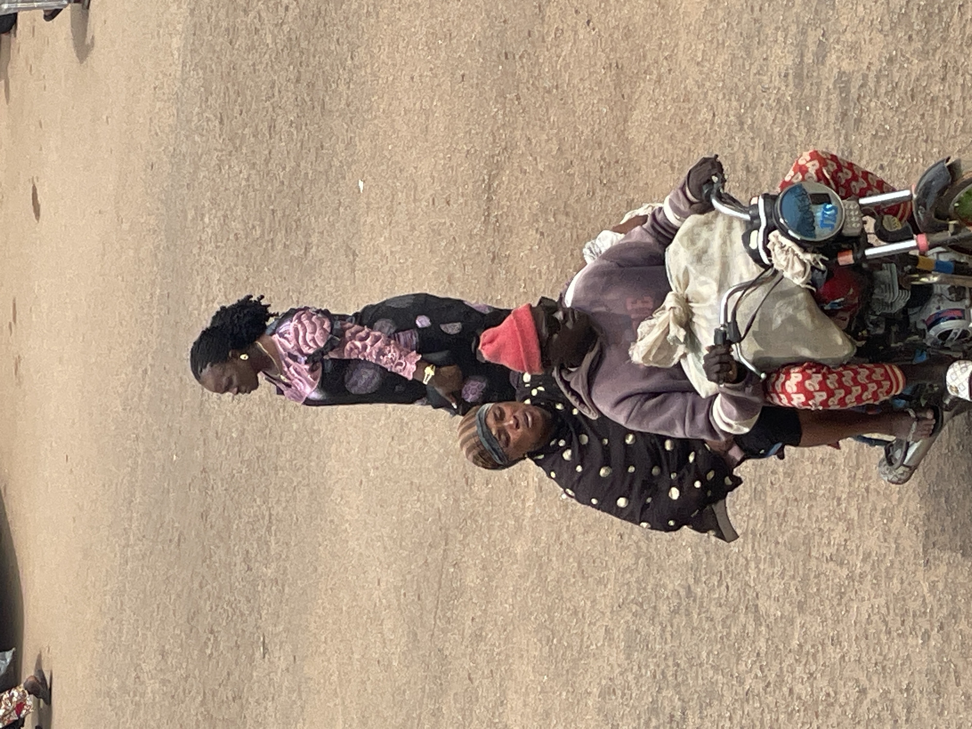A medium-angle, high-angle shot shows three people on a motorcycle in an urban environment. The motorcycle is dark colored with chrome accents. It is driven by a man wearing a purple hoodie, sunglasses, and a red beanie. He is carrying a large, light-colored bundle secured to the front of the motorcycle. He is wearing red and white patterned pants. Behind him, a woman is seated, wearing a black patterned headscarf and a black and white polka dot garment. Behind her, a woman in a black and pink patterned dress stands and appears to be looking to the side. The ground is a rough, sandy texture, typical of a dirt road or unpaved urban area.