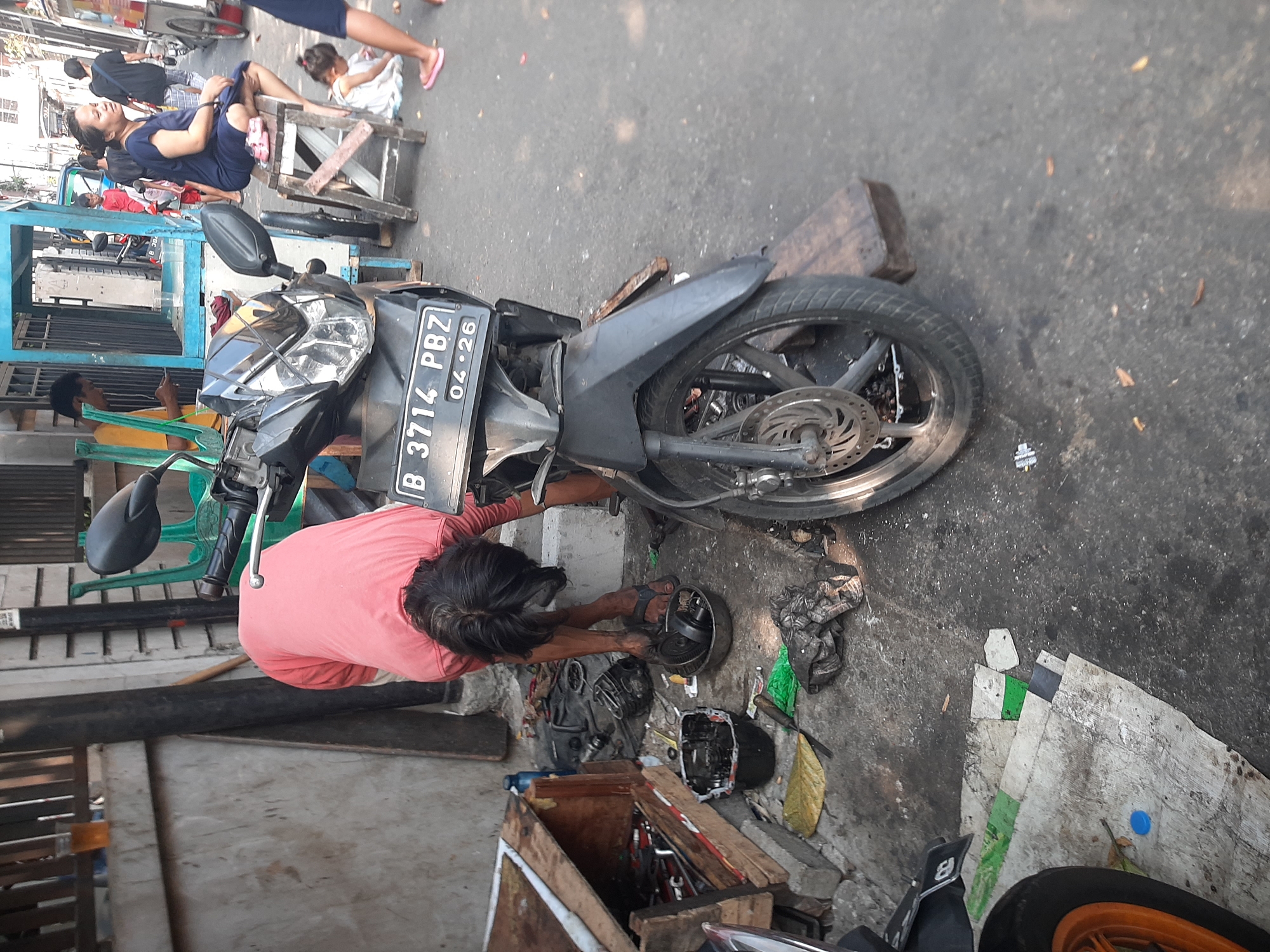 The image depicts an outdoor scene in a city environment, likely a street or alley in Central Jakarta, Indonesia, given the context provided. In the foreground, a person wearing a pink t-shirt is bent over a disassembled motorcycle, appearing to be in the process of repairing it. Various motorcycle parts, tools, and a toolbox are scattered around the ground. The motorcycle itself has a license plate that reads "B 3714 PBZ 04-26".
In the background, several other people are visible. A woman in a dark blue dress is sitting on a wooden stool. A young child in a white dress is on the ground near her. Other individuals are standing or walking in the background, with one person wearing a dark t-shirt and another in a red shirt. A blue stall or structure is present, suggesting a commercial area. The ground is paved with asphalt, and there are some wooden planks and concrete elements in the scene. The overall impression is that of a busy street, possibly a workshop or a roadside vendor area.