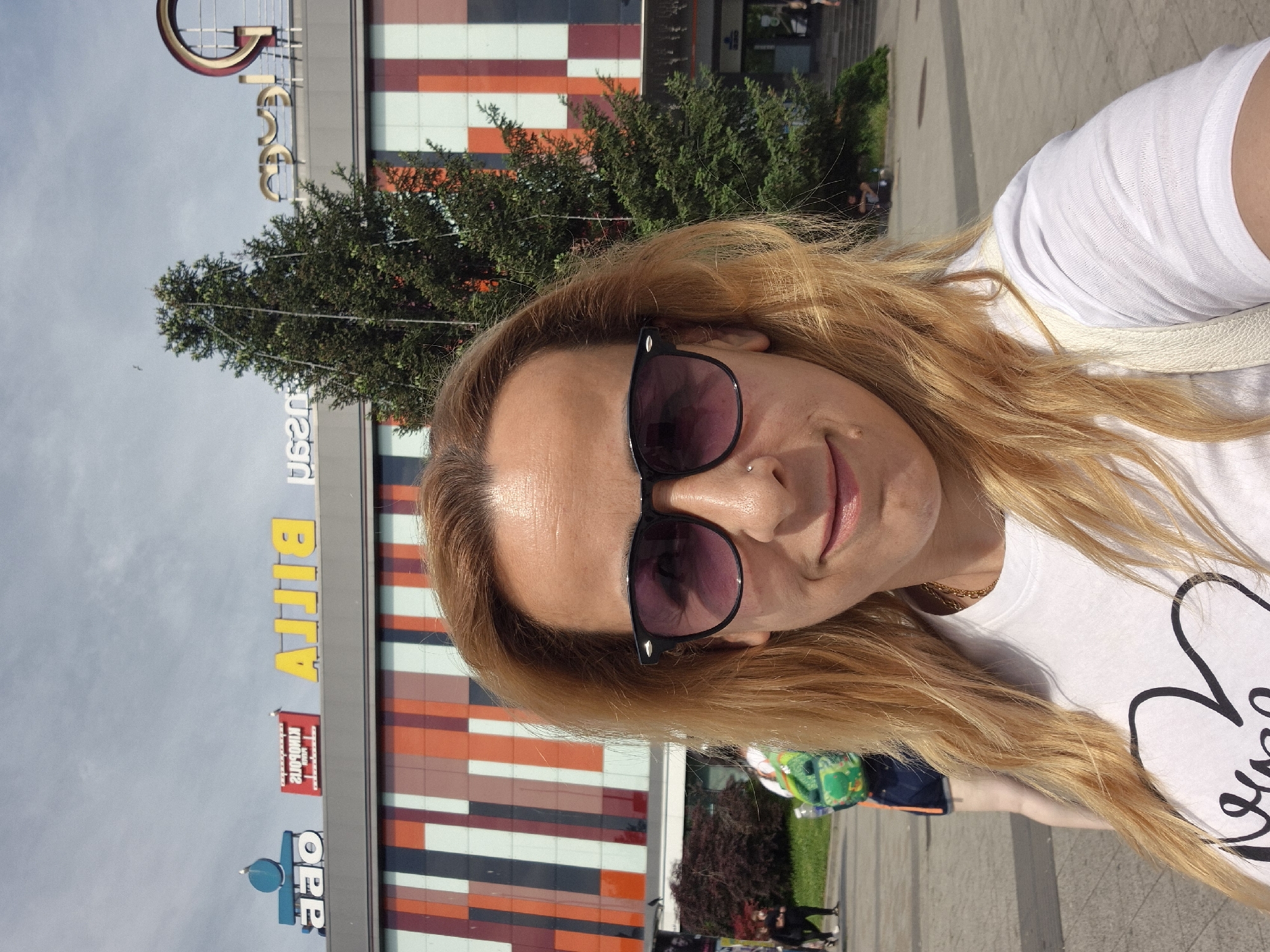 A woman in sunglasses is posing for a selfie in front of a building with colorful stripes. The building appears to be a shopping center, as there are storefront signs visible. The signs include "Oer" and "Billa." The background of the image shows trees, other pedestrians, and the sky.