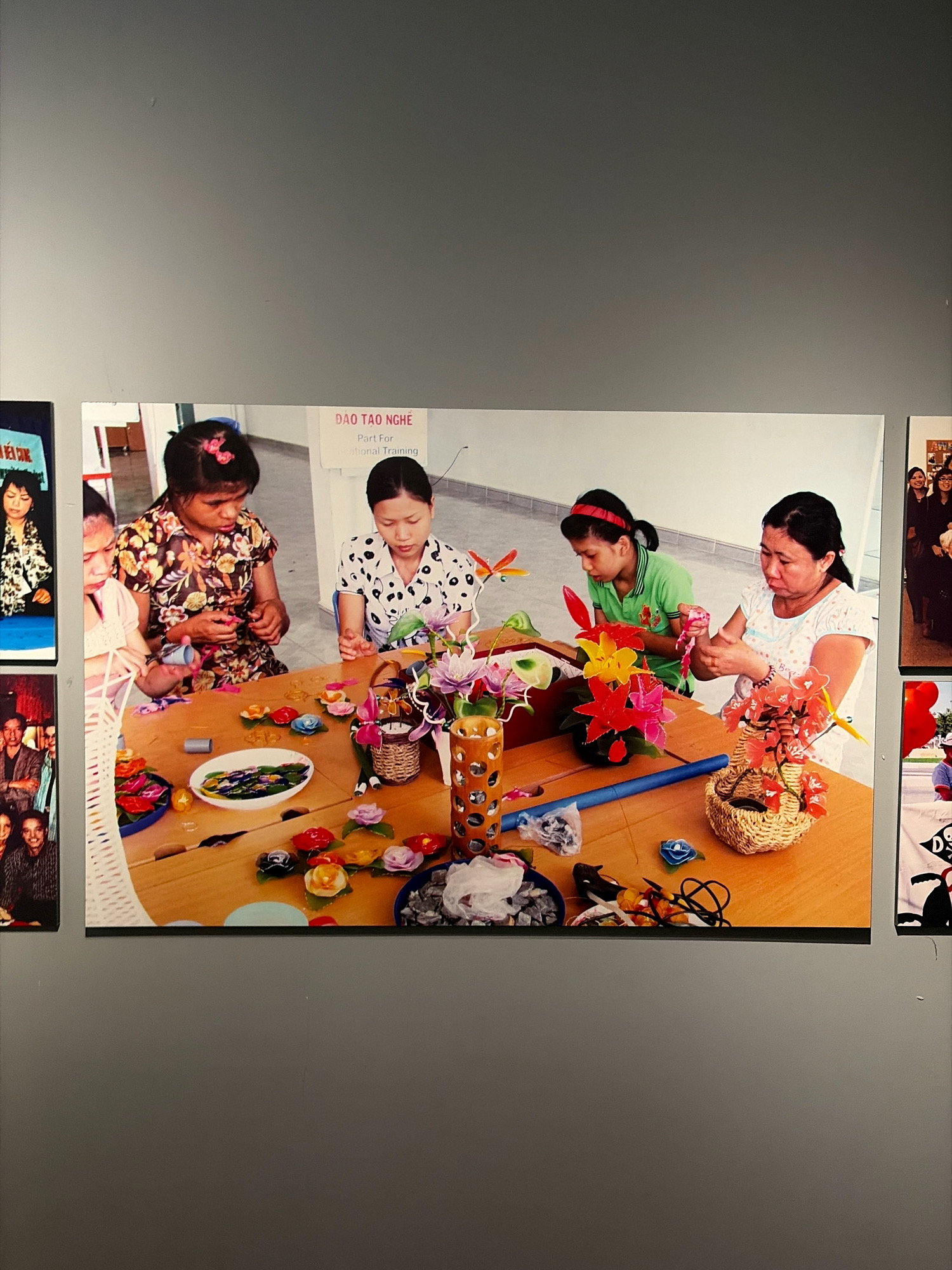 The image depicts four women sitting around a wooden table, engaged in a craft activity. The woman on the far left is partially visible, wearing a light-colored blouse and has dark hair. Next to her, a woman with dark hair and a floral print blouse is focusing on a task at the table. In the center, another woman with dark hair and a white blouse with black polka dots is also intently working. On the far right, a woman in a green shirt with a red headband is actively involved in crafting. The table is covered with colorful crafting supplies, including various flower-shaped objects, a woven basket with red flowers, and a white bowl containing what appear to be small stones. The background suggests an indoor setting with a white wall bearing a sign with Vietnamese text, "ĐÀO TẠO NGHỀ" which translates to "Vocational Training." The overall atmosphere appears to be one of shared activity and focus on creative work.