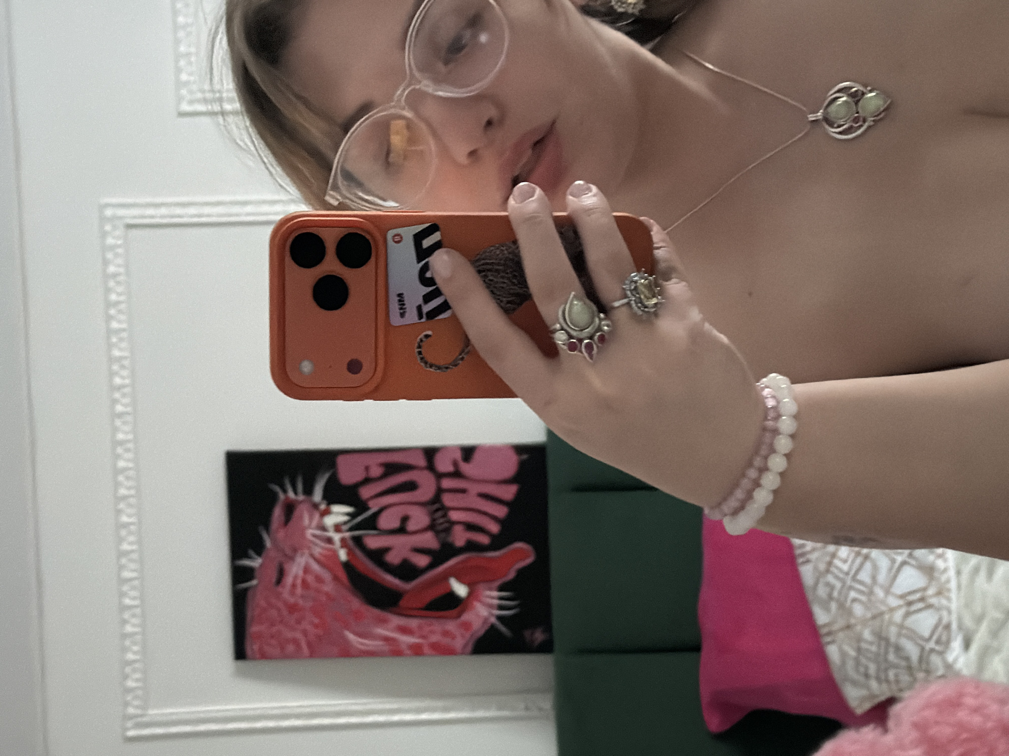 A selfie taken indoors shows a woman with light skin, wearing glasses and jewelry, including a necklace, bracelets, and rings. She is holding an orange smartphone with a sticker on the back, presumably to take the picture. Behind the phone and the woman's arm is a framed picture on a wall. The picture depicts a pink animal with the text "FUCK THIS SHIT" above it. The background includes a white wall with ornate trim, a dark green padded headboard, and pink and white cushions. The location is not discernible from the image.