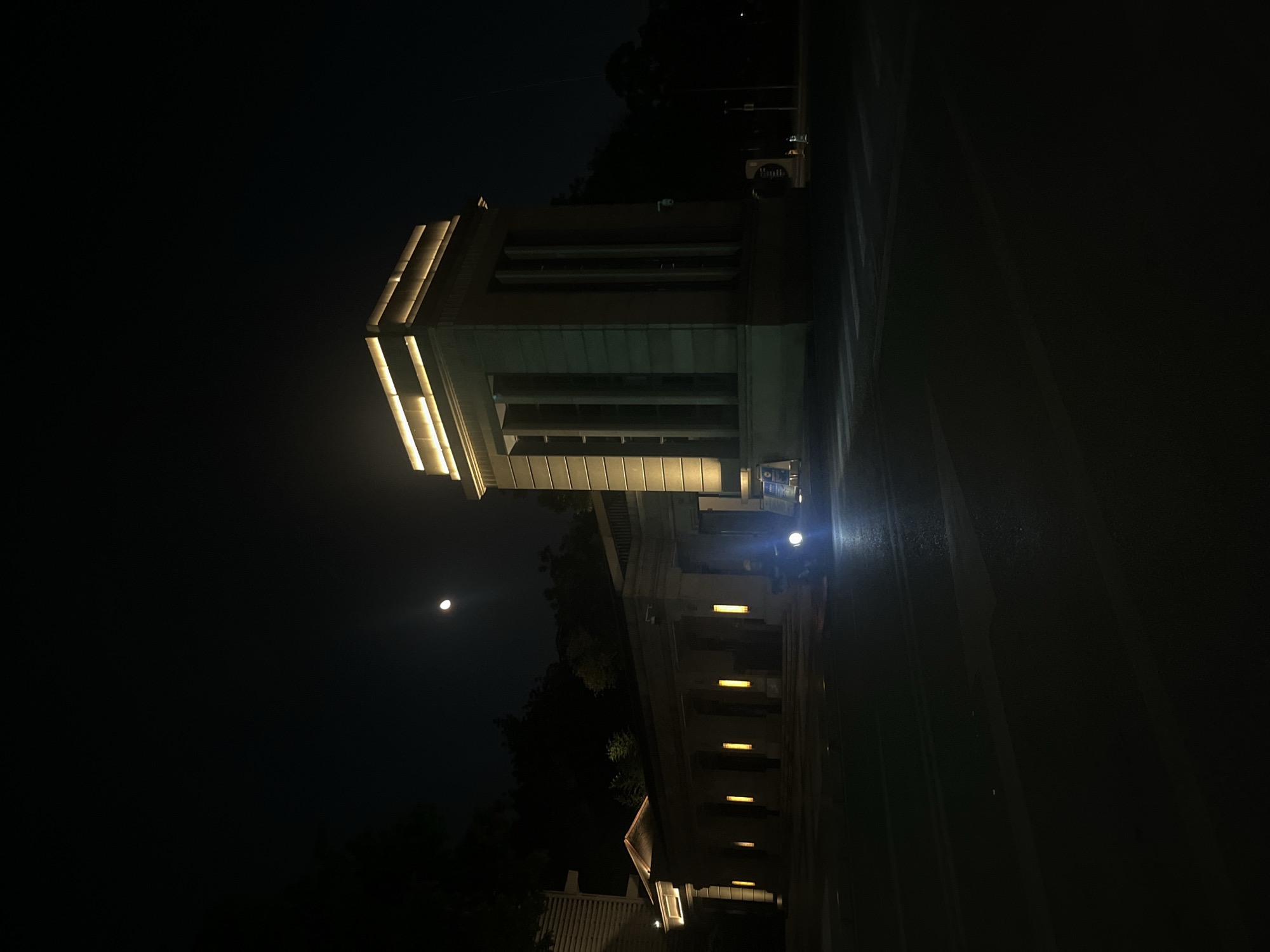 The image displays a nocturnal scene within Jiulongpo District, China. The central focus includes two distinct, illuminated architectural structures and a roadway with a two-wheeled vehicle.
On the right side of the frame, a prominent, light-colored building stands, characterized by a rectilinear form and vertical facade elements resembling pilasters or columns. The top section of this building is uniformly illuminated by horizontal strips of warm yellow light. Adjacent to its base, two small signs with blue upper sections and some orange traffic cones are discernible.
To the left and slightly receding, a lower, elongated building complex features a series of recessed openings or arches forming a colonnade or portico. This structure is vertically illuminated by warm yellow strip lights integrated into its facade. Dark trees and foliage are visible behind and to the left of this building, blending into the dark background.
The sky overhead is dark, indicating nighttime. A distinct crescent moon is visible in the upper left quadrant of the frame, emitting a soft glow.
In the foreground and midground, a dark asphalt roadway stretches across the scene. Faint white pedestrian crosswalk markings are present on the road surface. A two-wheeled vehicle, likely a scooter or motorcycle, is positioned on this roadway between the two buildings. It displays two bright, circular white headlights that intensely illuminate the road directly in front of it. A dark silhouette of a rider is visible above the vehicle's handlebars. The overall scene is defined by the contrast between the artificial warm yellow lighting of the buildings and the stark white light of the vehicle, set against the dark night and the natural moonlight.