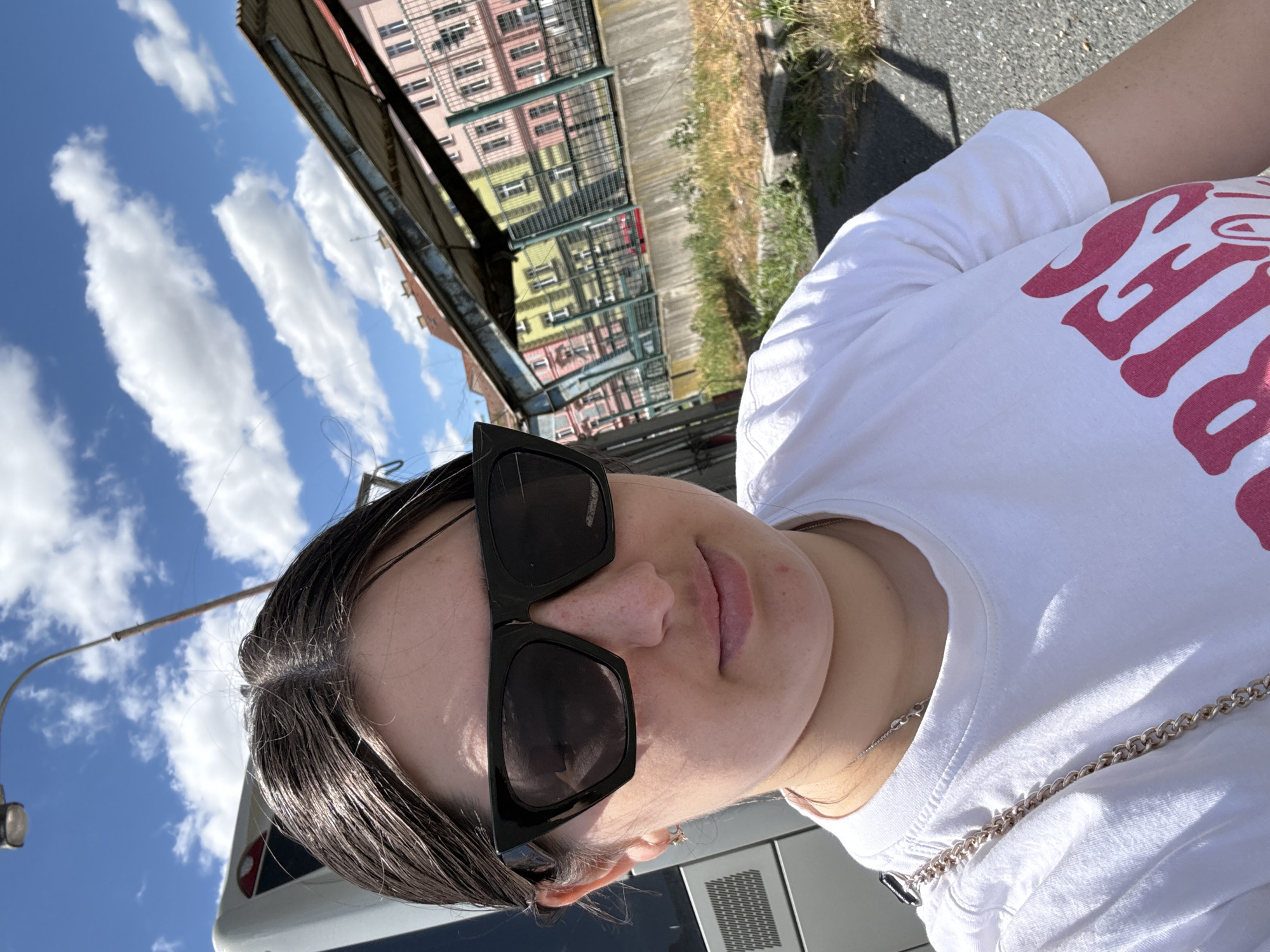 A young woman with dark hair, wearing large black sunglasses and a white t-shirt with pink lettering, is taking a selfie outdoors. She is positioned in the foreground, with a bright blue sky and white clouds behind her. To the right, a row of colorful buildings in shades of pink, yellow, and green is visible, with a metal structure in front of them. The woman has a chain strap bag draped over her shoulder. The background suggests an urban environment, possibly a public space or street.