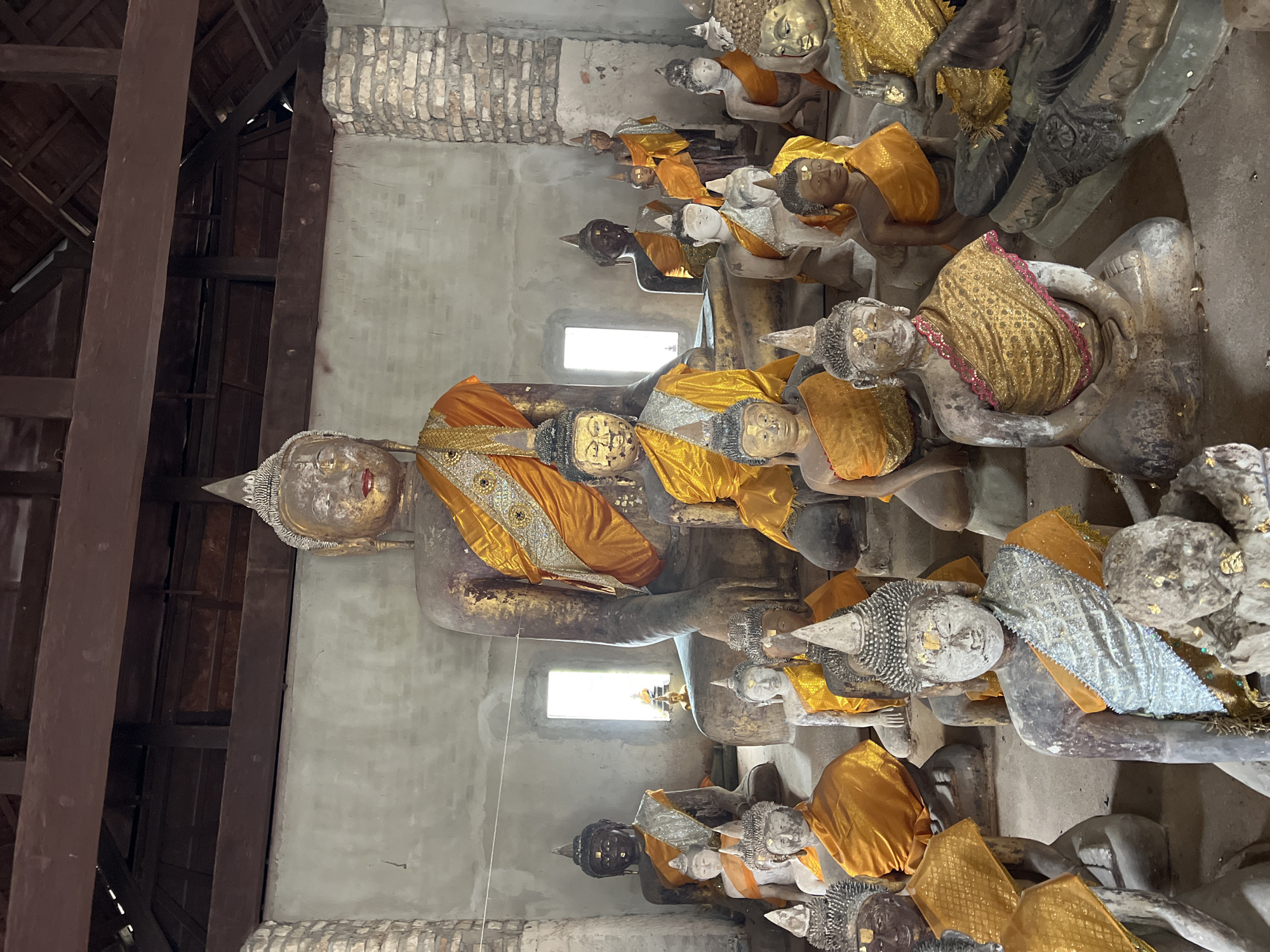 This image shows a collection of Buddha statues inside a building, likely a temple, in the Ko Samui District of Thailand. The statues vary in size and material, with some appearing to be made of stone and others of bronze or wood. Many of the statues are adorned with draped saffron-colored cloth, a common practice in Buddhist traditions. The building itself appears old, with exposed wooden beams and brickwork visible. The lighting suggests an indoor environment, possibly with natural light filtering in from windows. The overall scene conveys a sense of religious devotion and historical significance.