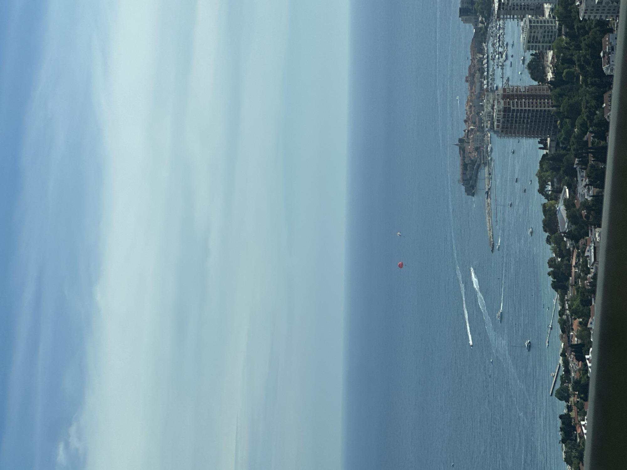 The image is an aerial view of a coastal city, likely Vrela, Montenegro, on a clear day with scattered clouds. The deep blue sea occupies the majority of the frame, with the horizon clearly defined. Several boats are visible on the water, some leaving white wakes, indicating movement. In the distance, a red parachute is visible, suggesting recreational activity. The coastline is lined with buildings, including residential areas and a prominent harbor filled with yachts and smaller vessels. A historic walled town with red-tiled roofs and a church spire is situated on a peninsula jutting into the sea. In the foreground, modern buildings, including a high-rise under construction, are visible alongside lush green trees. A dark railing runs along the very bottom edge of the frame, suggesting the photo was taken from a high vantage point.