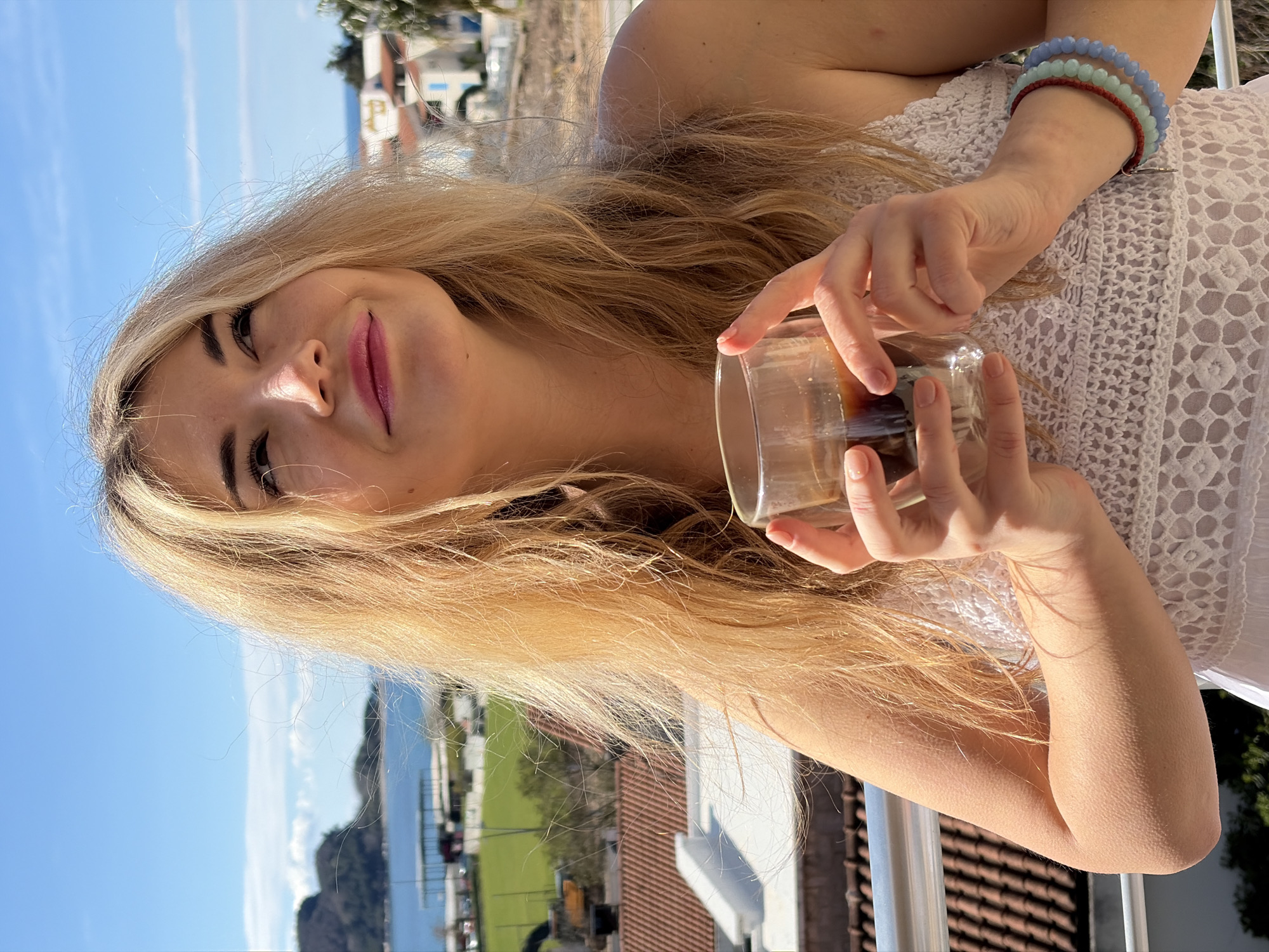 A blonde woman, wearing a white lace top, is photographed from a low angle against a bright blue sky and a cityscape featuring terracotta rooftops and coastal scenery. She holds a glass containing a dark liquid, possibly coffee or tea, with her fingers visible around the rim. A stack of bracelets adorns her wrist. The scene likely overlooks the city of Budva, Montenegro, recognizable by the buildings and coastal views.