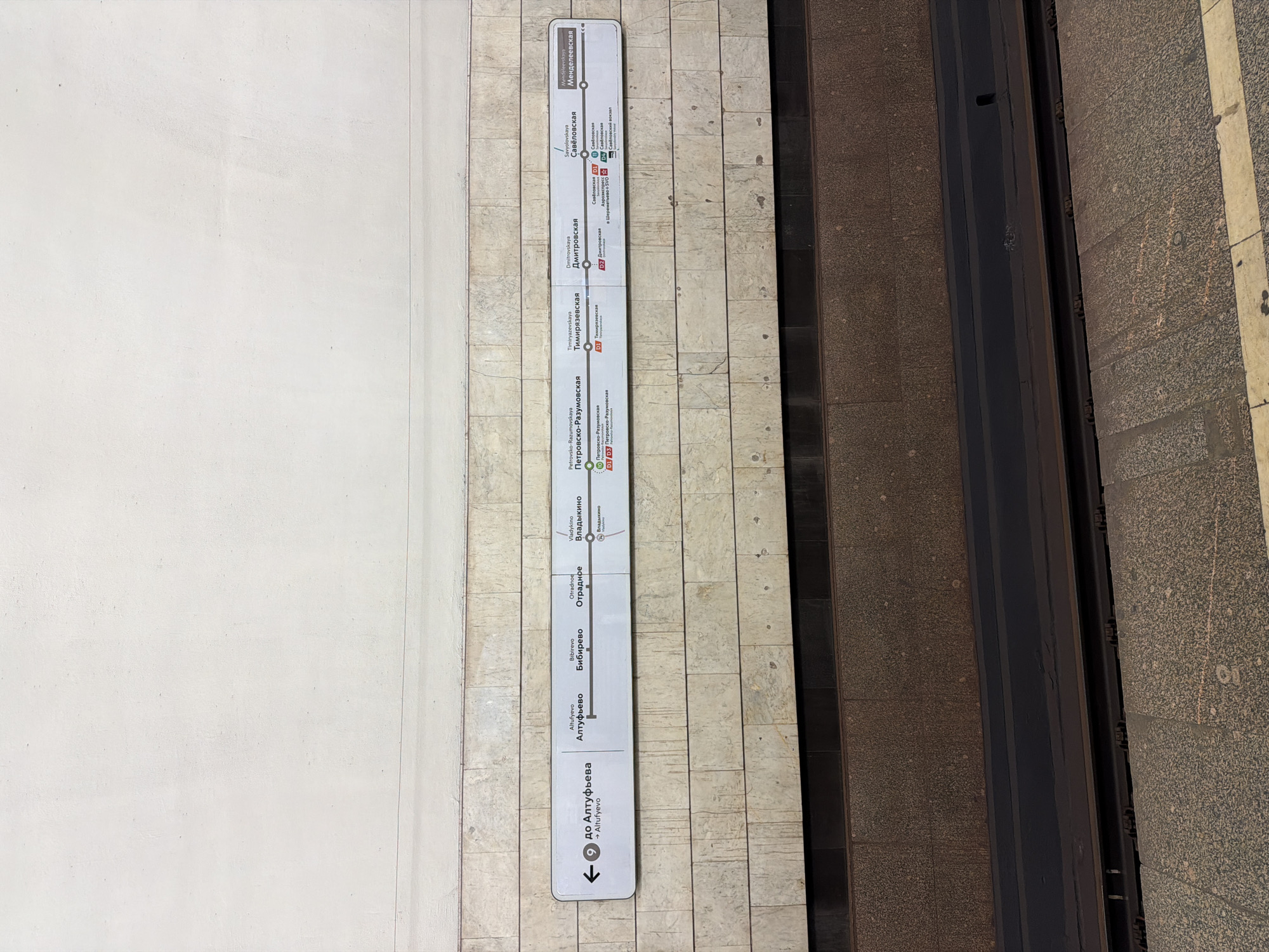 The image depicts a vertical sign showing the Moscow Metro Line 9, located in Moscow, Russia. The sign, affixed to a wall, lists the stations, from "Altufyevo" at the bottom, progressing upwards through "Bibirevo," "Otradnoe," "Vladykino," "Petrovsko-Razumovskaya," "Timiryazevskaya," "Dmitrovskaya," "Savyolovskaya," and ending with "Mendeleevskaya" at the top. The direction to "Altufyevo" is indicated with an arrow at the bottom. There are symbols indicating transfers to other metro lines and the Moscow Central Diameters (MCD) at certain stations. The sign is adjacent to the edge of a train platform, including the platform edge and the train itself.