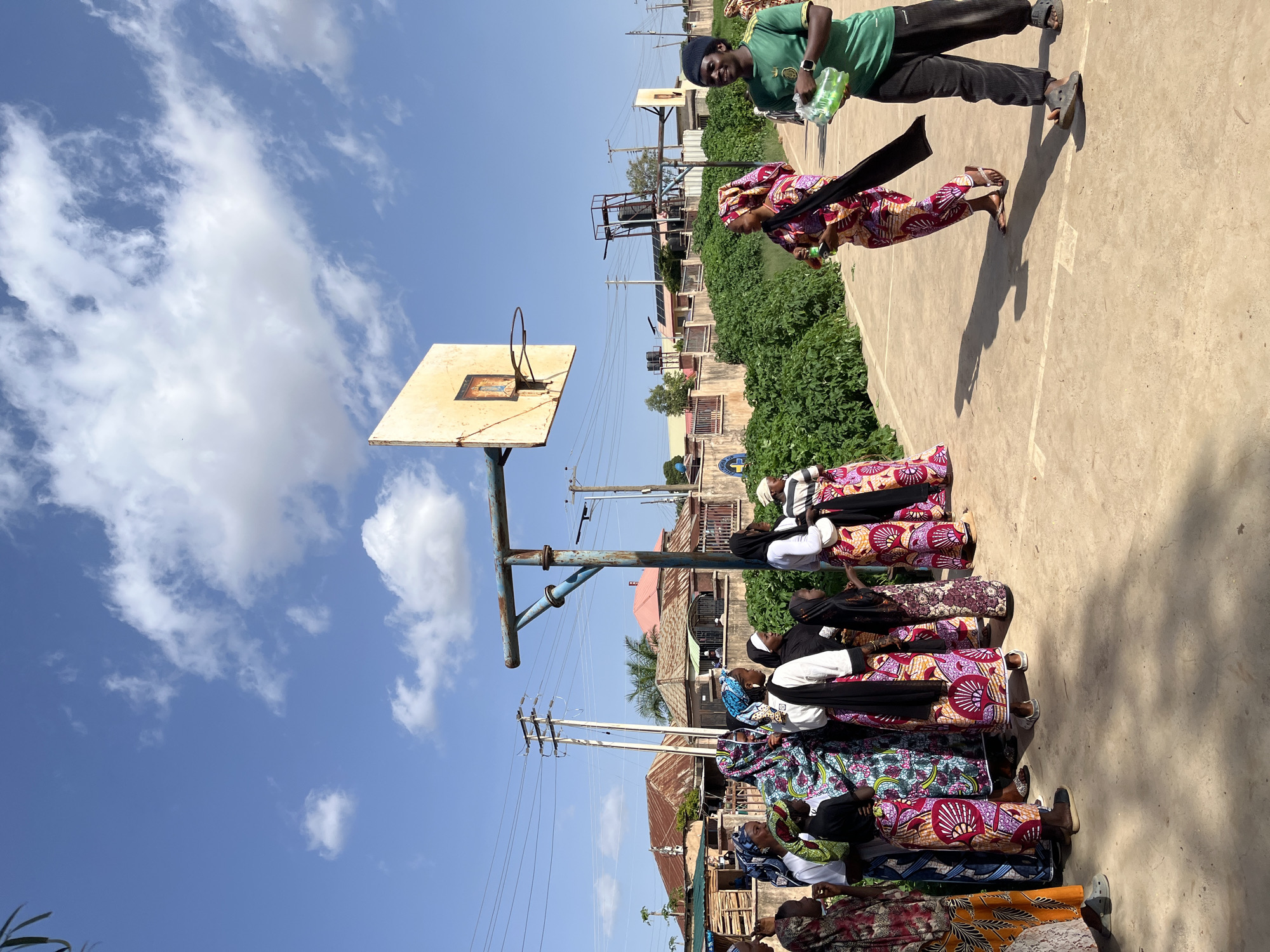 In Gura Top, Nigeria, a group of people are gathered on a basketball court under a clear blue sky with scattered clouds. The court appears worn, with visible lines and patches of dirt. A rusty blue basketball hoop with a faded backboard stands prominently on the left side of the frame.
On the left, a cluster of individuals, mostly women and children, are dressed in colorful patterned clothing and headscarves. They stand in a line, facing towards the center of the court. Their postures suggest they are observing an activity or waiting.
On the right side of the court, two individuals are in motion. A woman in a vibrant, patterned dress and headscarf is walking away from the viewer, holding a green bottle in her right hand. To her right, a young man in a green t-shirt and dark pants is walking towards the viewer, carrying a plastic bag filled with several green bottles. His attention is directed towards the woman.
In the background, residential buildings with corrugated metal roofs are visible, interspersed with lush green vegetation and trees. Electrical poles and wires are also present, indicating a developed area. The overall scene depicts a casual gathering or event taking place outdoors on a sunny day.
