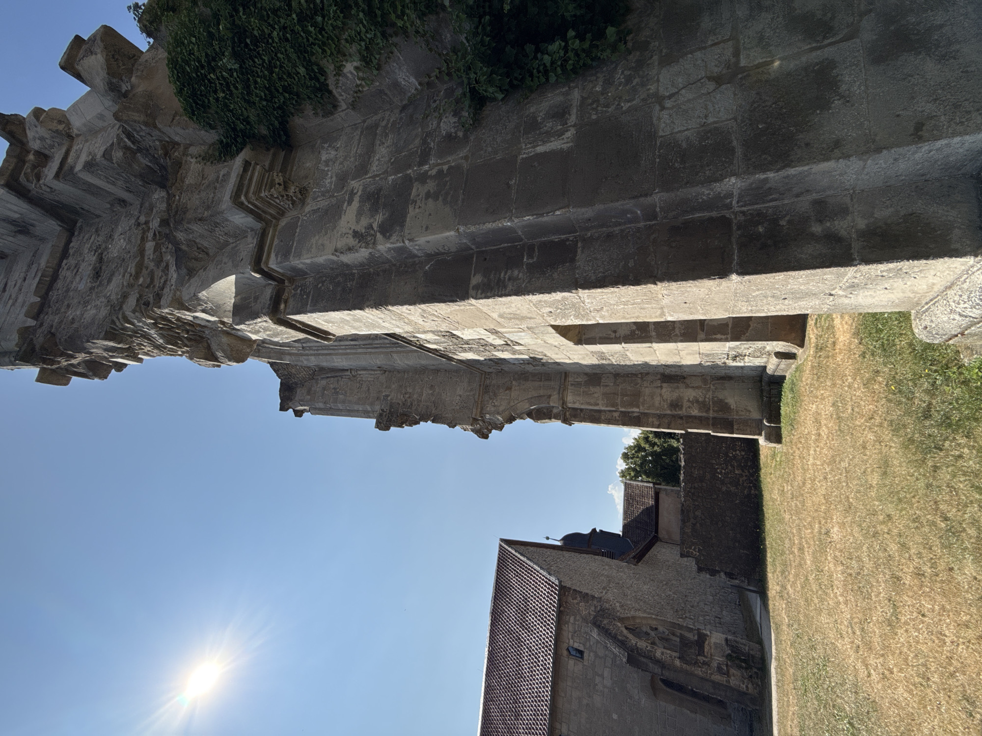 The image depicts the ruins of the Walkenried Abbey in Walkenried, Germany. The foreground is dominated by a section of the abbey's stone ruins, characterized by weathered blocks and intricate architectural details. Ivy climbs the side of the structure. To the left, a portion of a well-preserved building with a red-tiled roof and stone walls is visible, suggesting a functional part of the abbey complex or a nearby structure. The sky is a clear, bright blue with the sun prominently visible in the upper left quadrant, casting strong light and shadows. The ground is covered with dry, yellowed grass. The overall scene suggests a historical site on a sunny day.