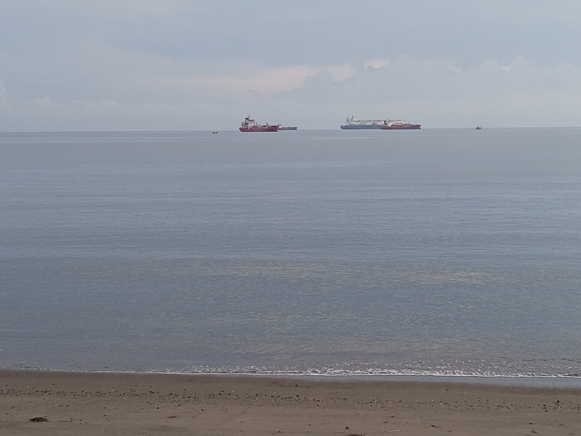 The image depicts a serene coastal scene in Lhokseumawe, Indonesia. The foreground features a sandy beach with gentle waves lapping the shore. In the distance, the calm sea stretches towards the horizon under a cloudy sky. Several ships are visible on the water, with at least two appearing to be tankers. One is red and white and the other is white and blue. There are also smaller fishing boats scattered on the water.
