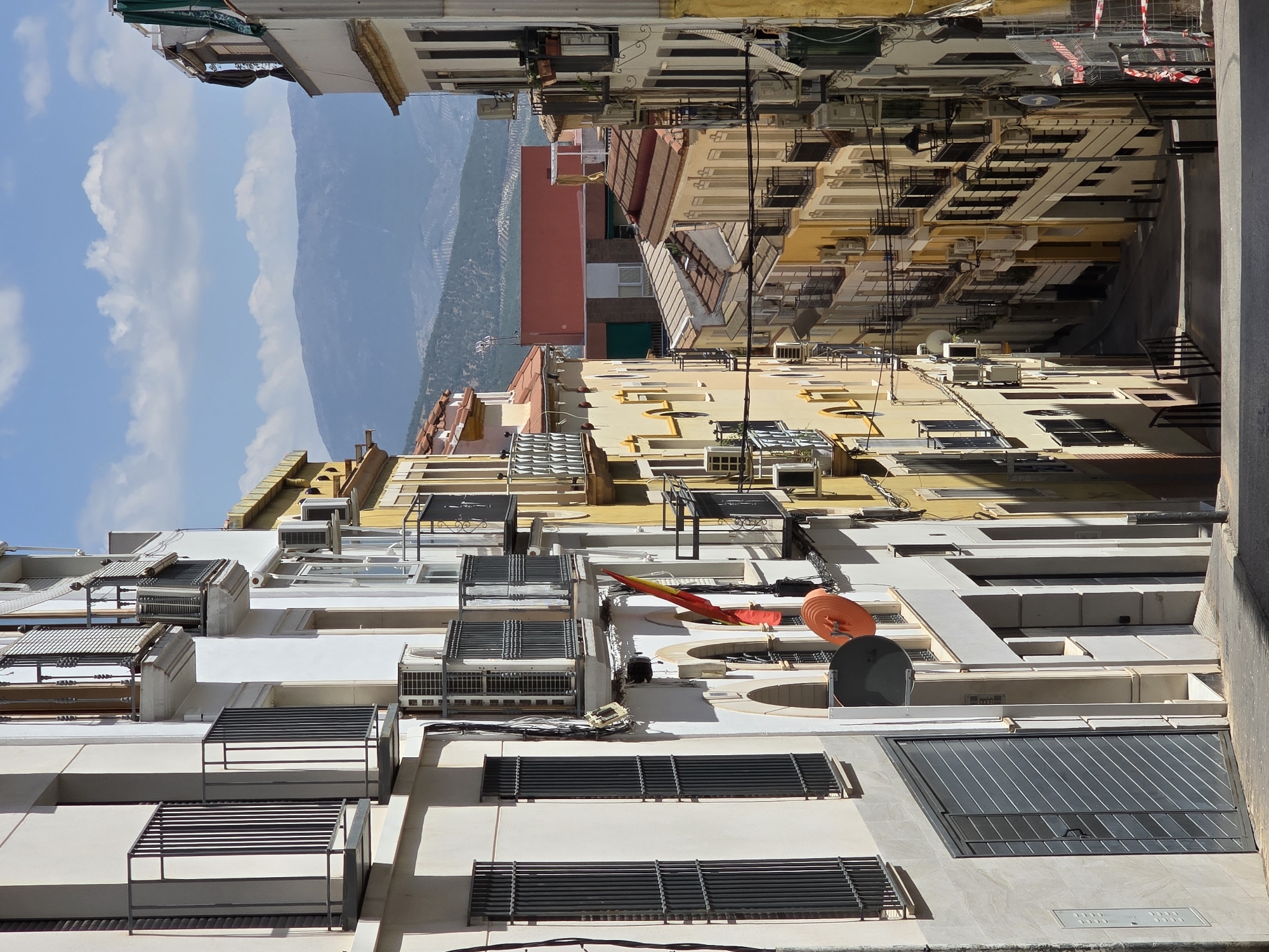 The image depicts a narrow, downhill street in Jaén, Spain, flanked by multi-story buildings. The architecture suggests a historic urban setting, with balconies, window shutters, and ornate building facades. On the left, a building with a light-colored facade features dark shutters and an industrial-style metal door. Further down, a Spanish flag is visible hanging from a balcony. The right side of the street is lined with buildings painted in various shades of yellow and cream, also adorned with balconies. Air conditioning units are visible on many of the buildings, indicating a warm climate. The street itself is paved with cobblestones or similar material, and it descends into the distance, where it curves out of view. In the background, distant mountains are visible under a partly cloudy sky. There are no people or animals visible in the image. The scene conveys a sense of everyday urban life in a Spanish city, characterized by its architecture and environmental setting.